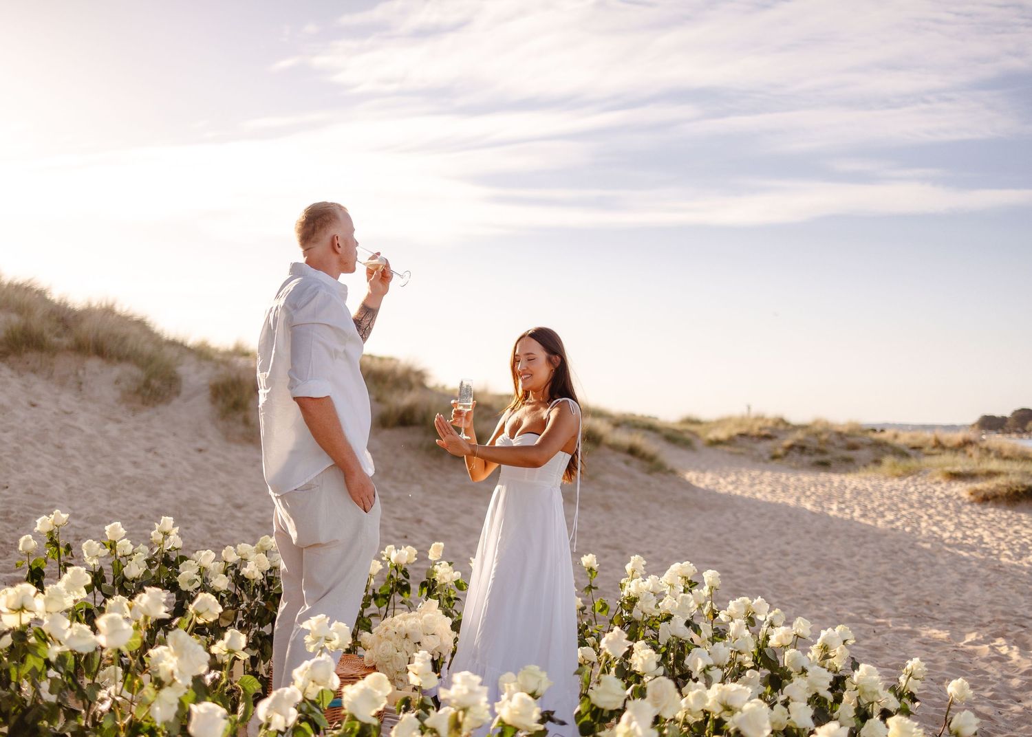Couple celebrating on a sandy beach surrounded by white roses at sunset, woman in blue dress, man in white suit.
