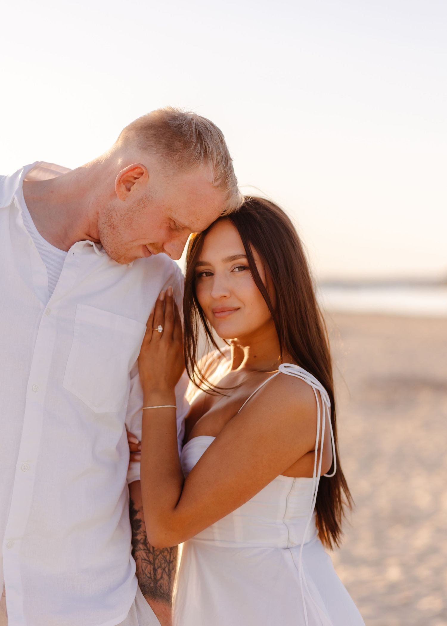 Romantic couple in white embracing on beach at sunset, woman smiling at camera.