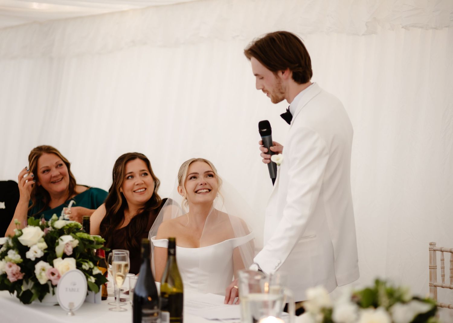 Groom in white suit giving speech at wedding reception as bride smiles joyfully at decorated table.