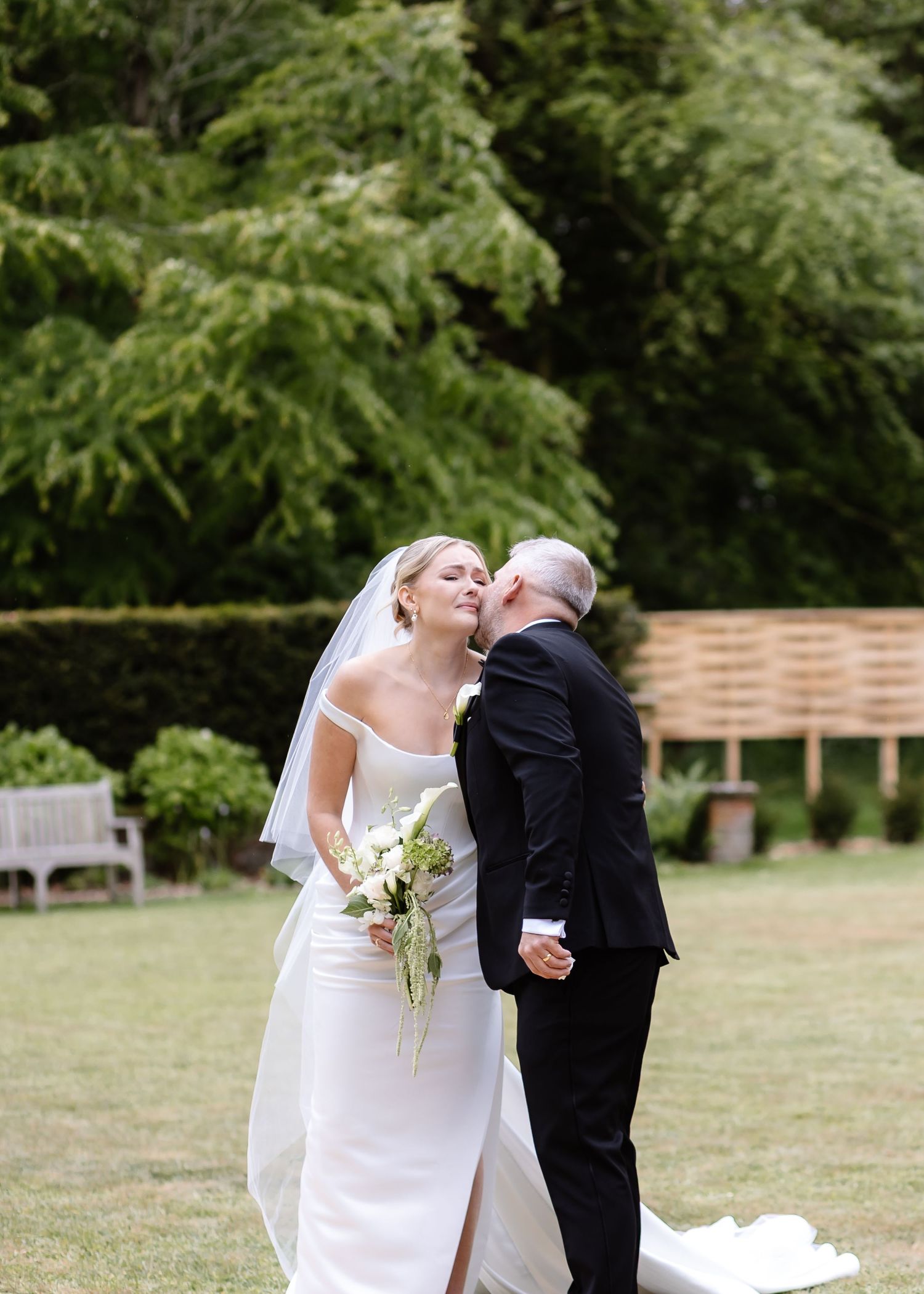 Bride in white off-shoulder gown with veil holds bouquet as groom in black suit kisses her cheek outdoors.
