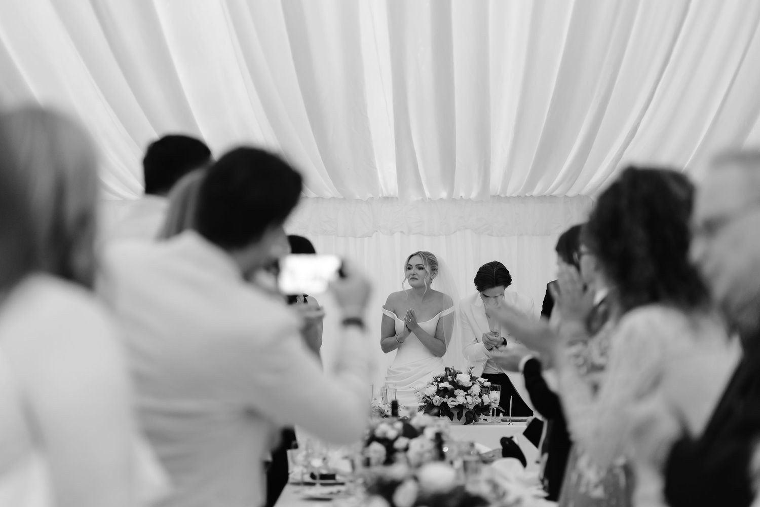 Black and white wedding reception photo of bride clapping at head table surrounded by guests under draped ceiling.