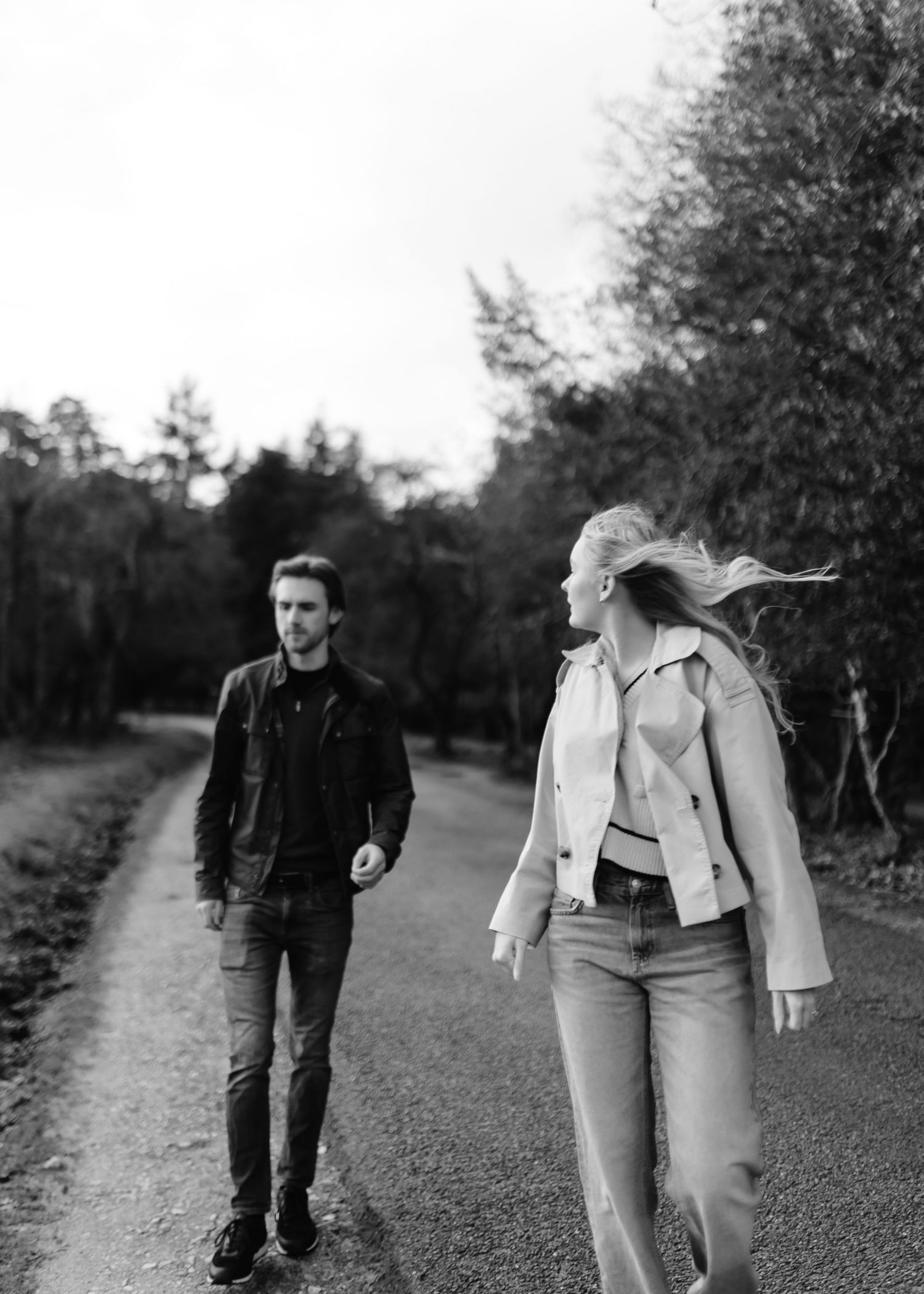 Black and white photo of a man and woman walking on a tree-lined road, woman's hair blowing in the wind.