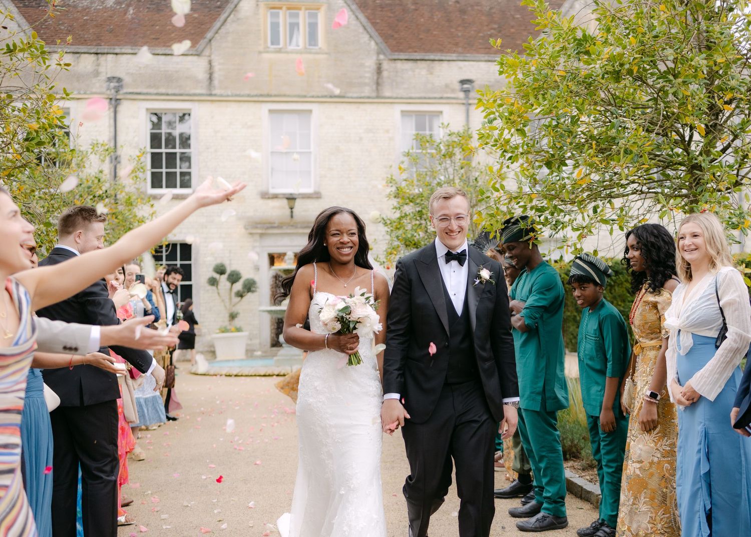 Bride and groom walk through confetti shower outside elegant stone manor, guests celebrating in colorful attire.