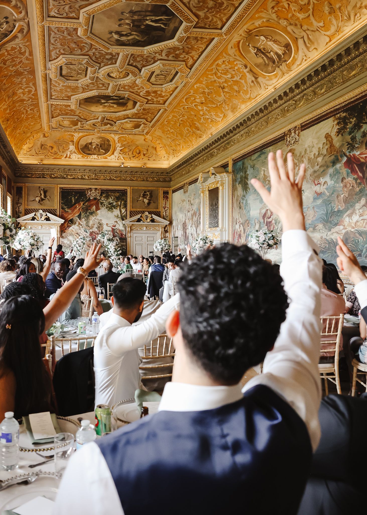 People with raised hands participate in a celebration inside an ornate room with gilded ceiling and frescoed walls.
