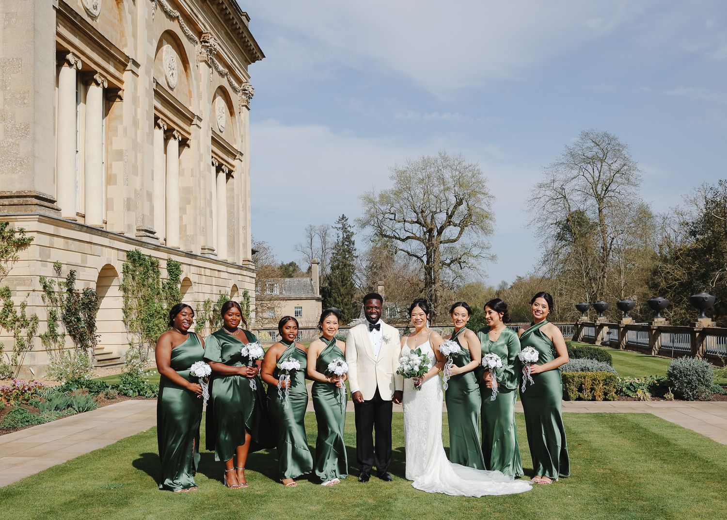 Wedding party poses outdoors in green bridesmaid dresses against a grand stone building on a sunny day.