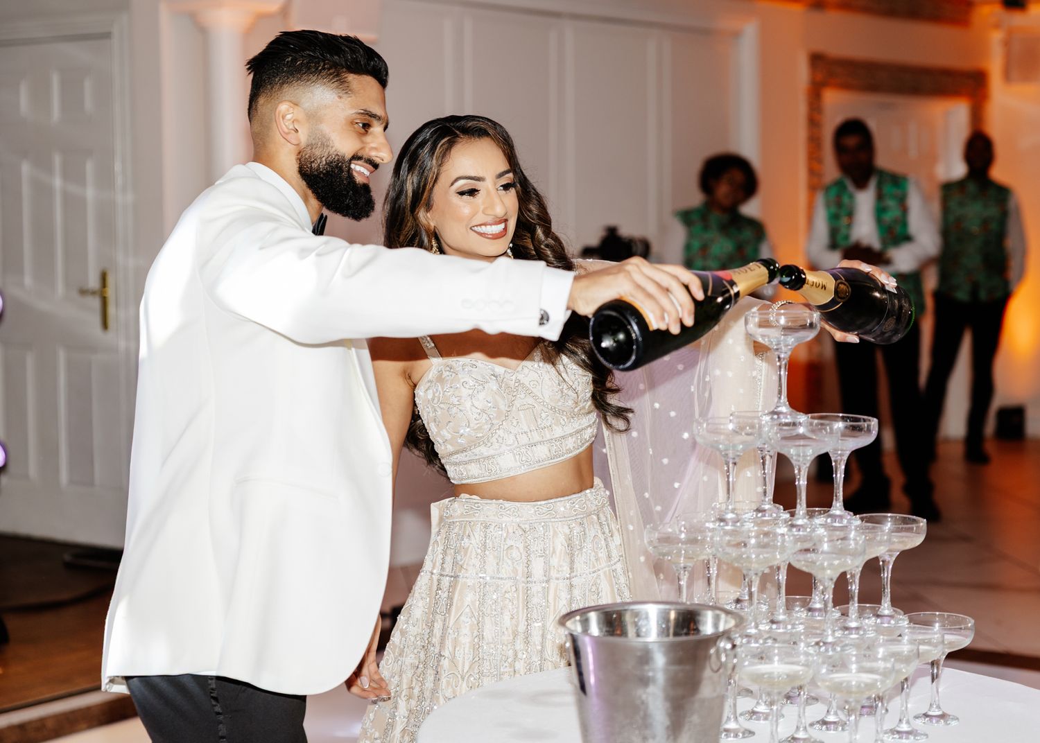 Wedding guests pour champagne into a tower of crystal glasses at an elegant reception celebration.