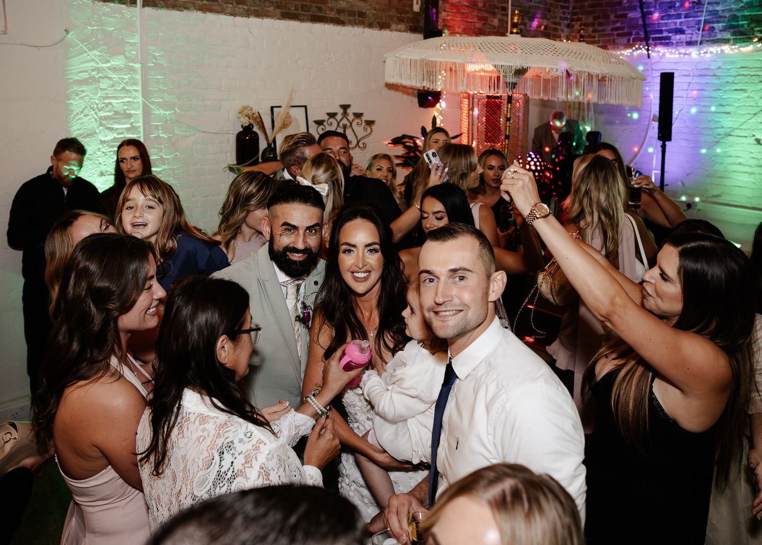 Wedding guests dance and celebrate on a crowded dance floor illuminated by colorful lights at a reception party.