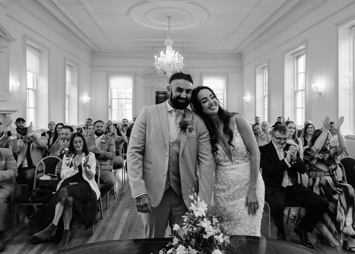Wedding guests celebrate joyfully in an elegant white ballroom with chandelier and large windows.