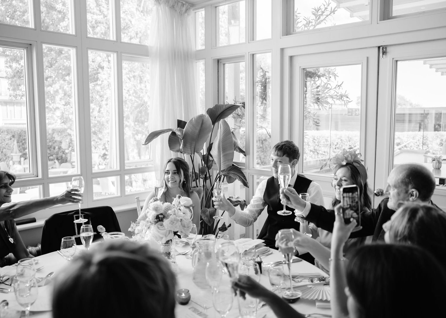 Black and white wedding reception toast with guests raising champagne glasses in elegant windowed dining room.