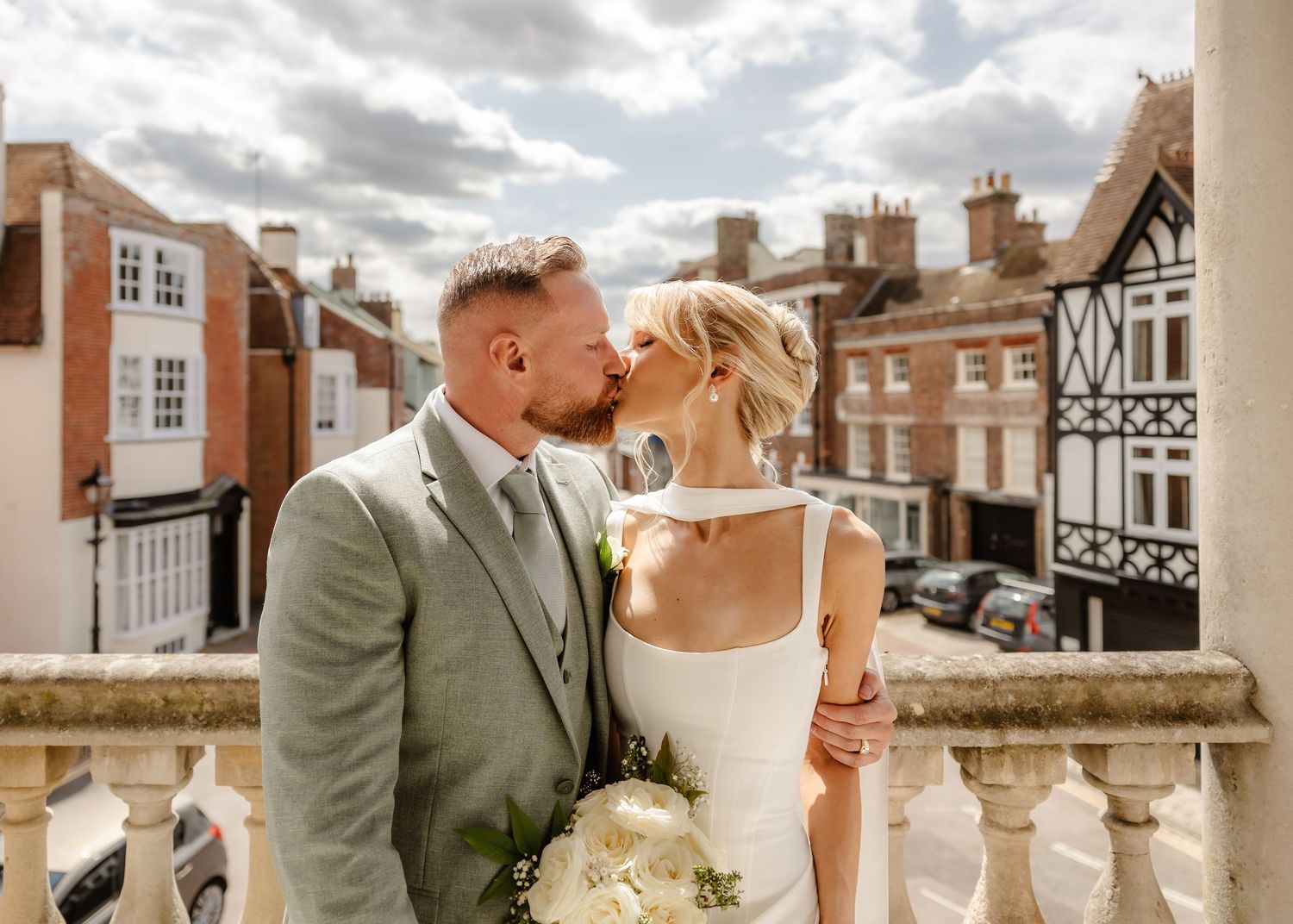Bride and groom share romantic kiss on balcony overlooking charming town street on wedding day.