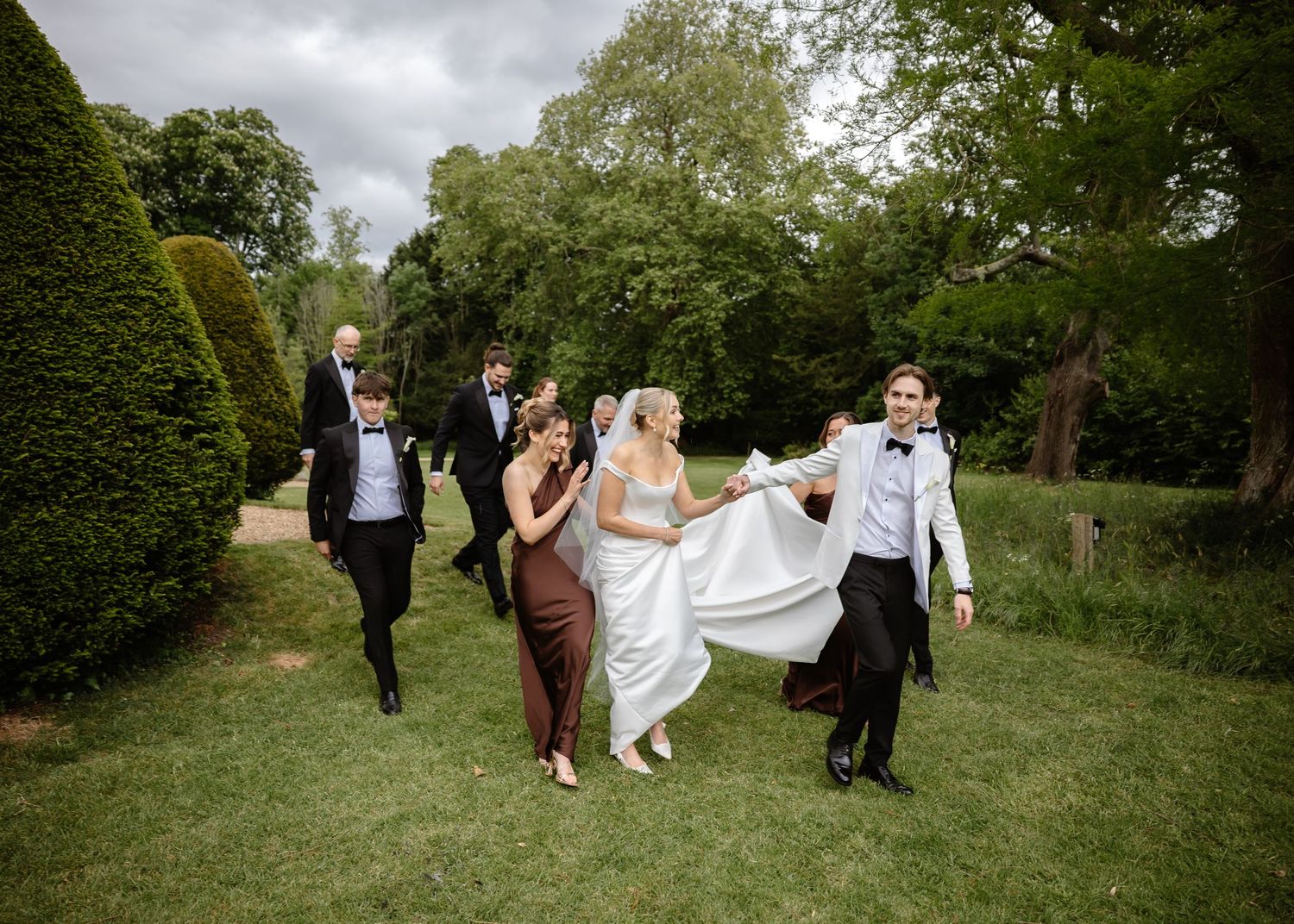Bride in white gown walks with wedding party across lush green garden, groomsmen in tuxedos and bridesmaids in elegant attire.