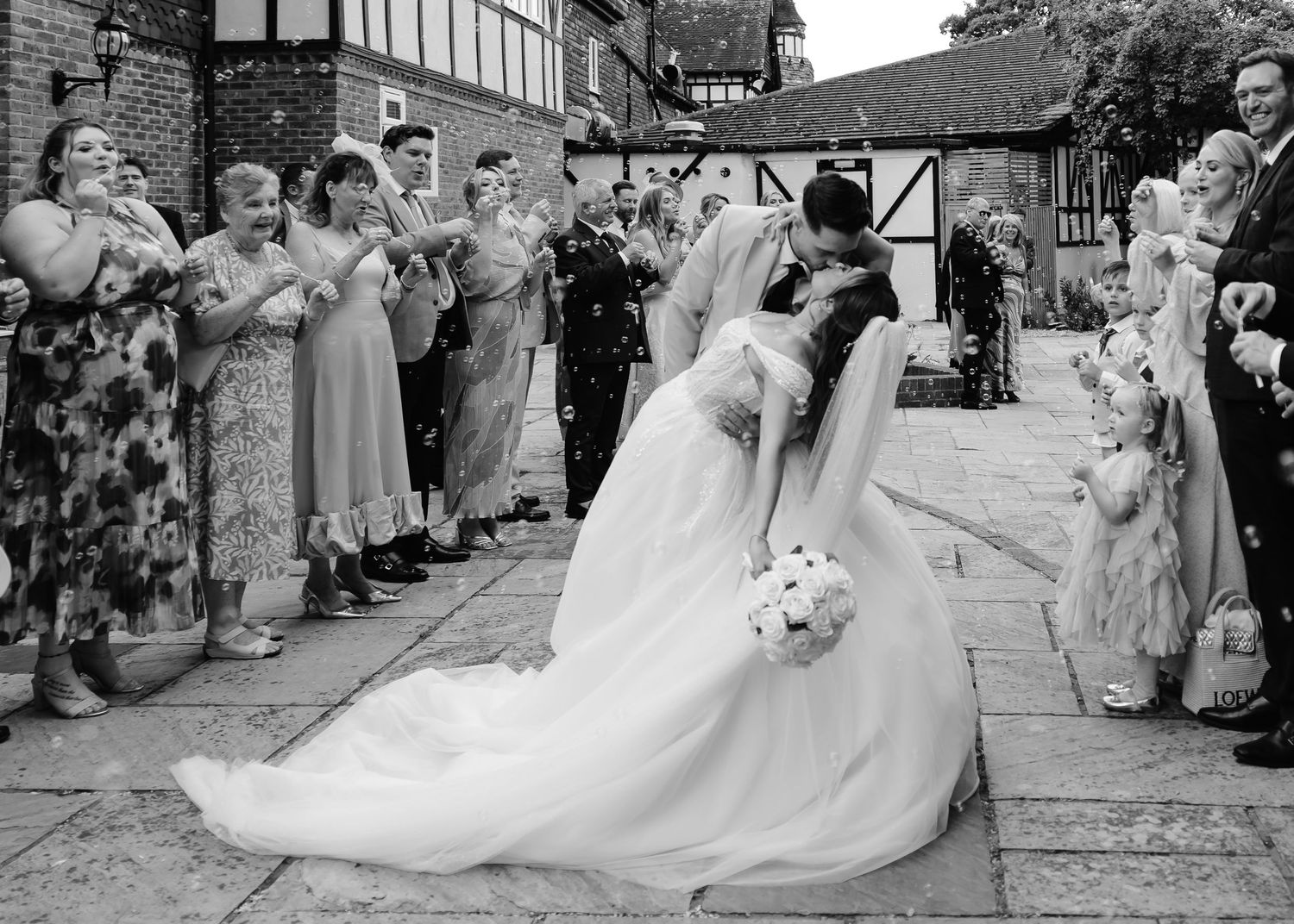 Black and white wedding photo of bride and groom kissing while guests throw confetti outside a venue.