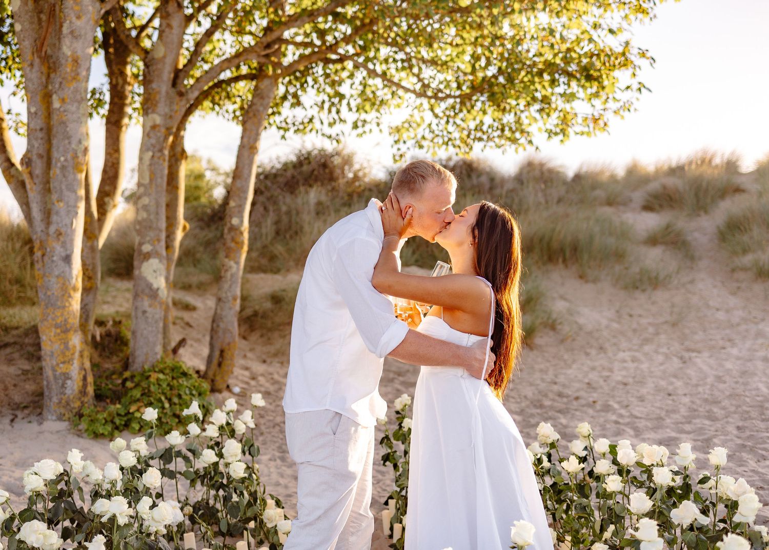 Couple sharing a romantic kiss at a beach wedding ceremony surrounded by white roses and golden sunset light.