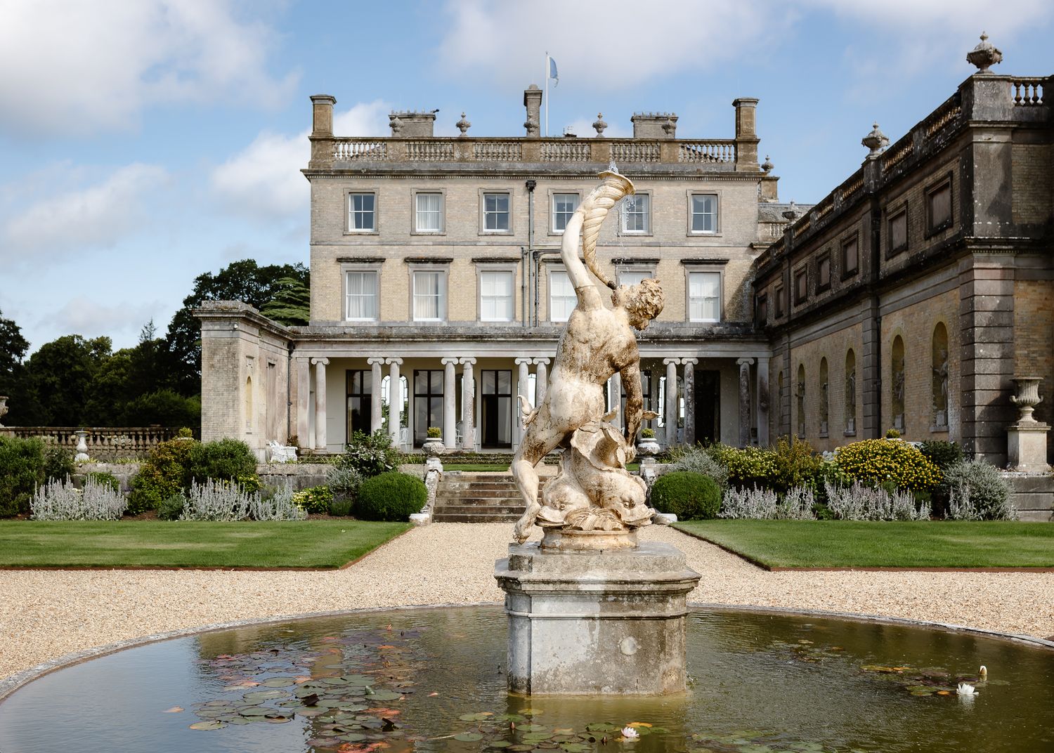 Elegant Georgian country house with ornate fountain statue and formal gardens under a partly cloudy sky.