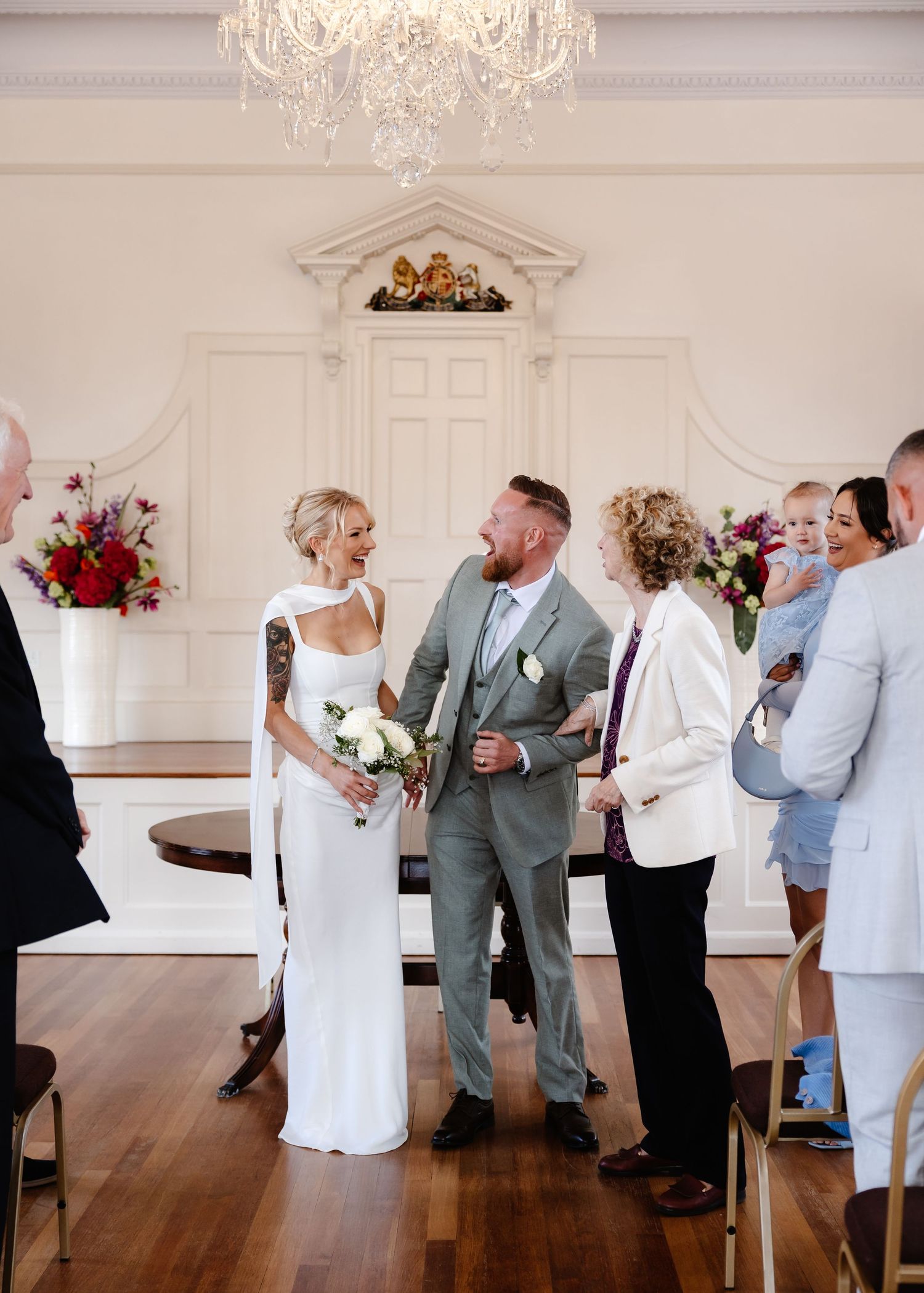 Bride and groom laughing together after wedding ceremony in elegant white venue with chandelier and floral arrangements.