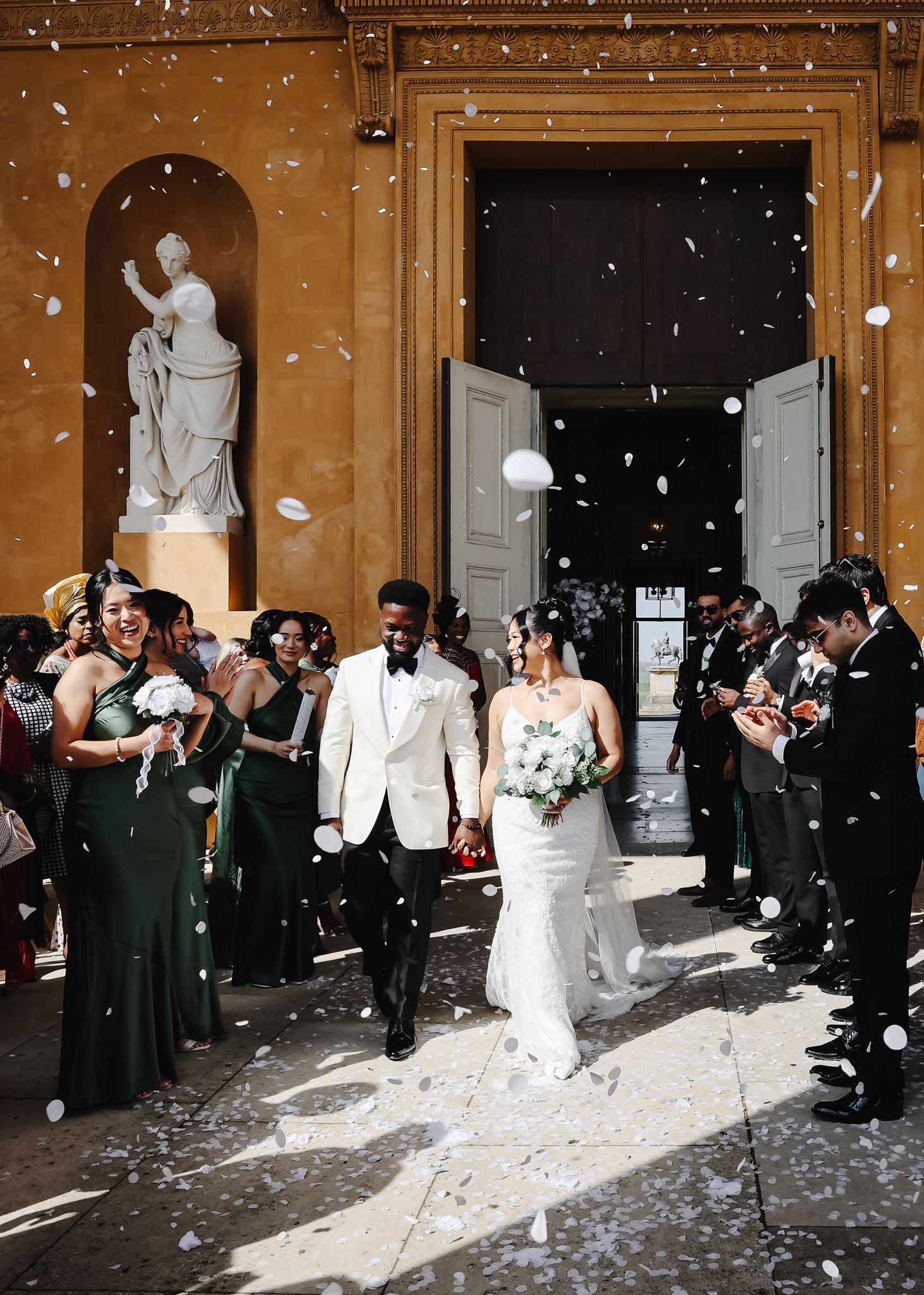 Newlywed couple exits elegant venue as wedding party throws white confetti, bride in fitted gown, groom in white tuxedo.
