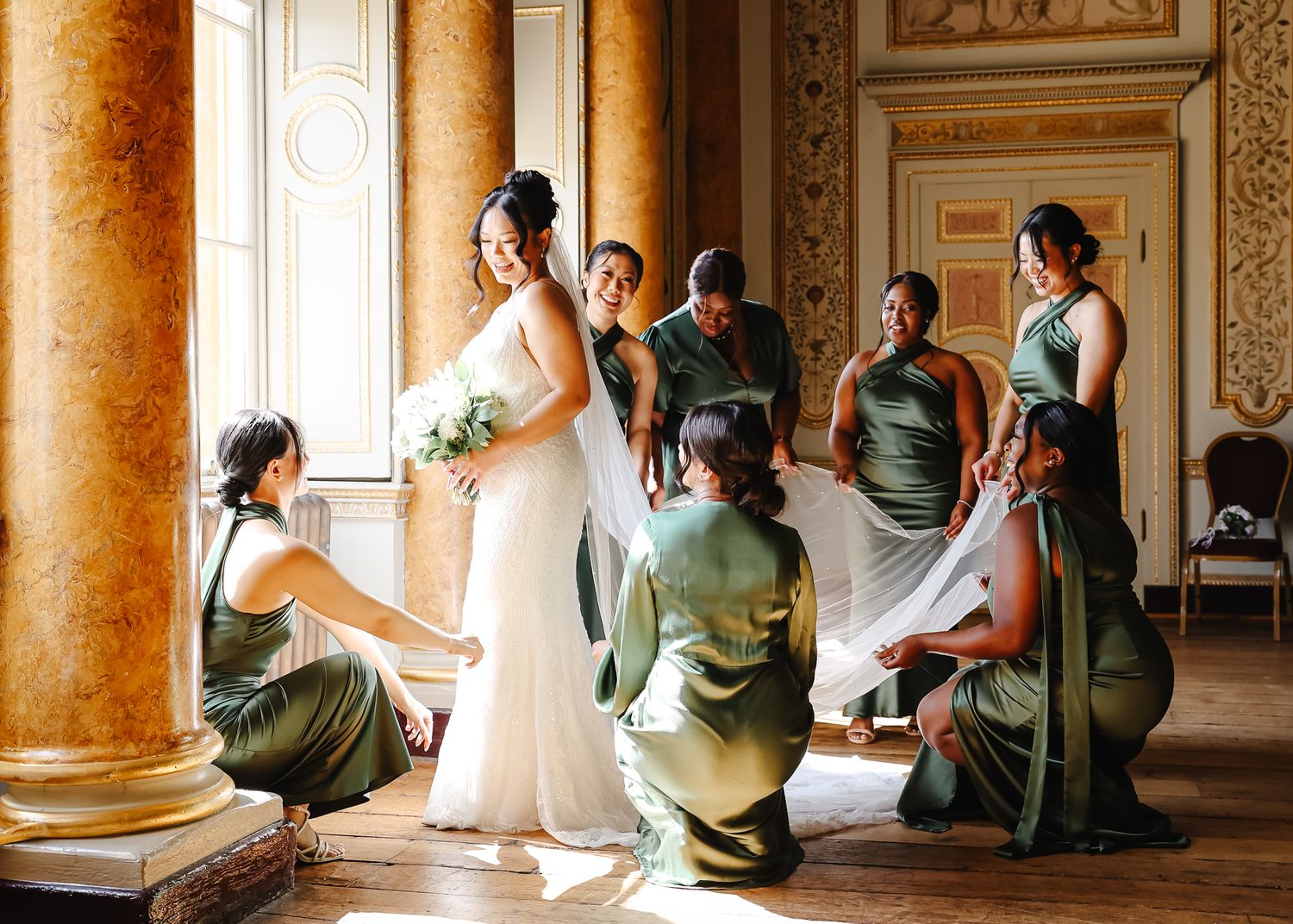 Wedding party in sage green dresses poses elegantly in a grand ballroom with ornate columns and natural light.