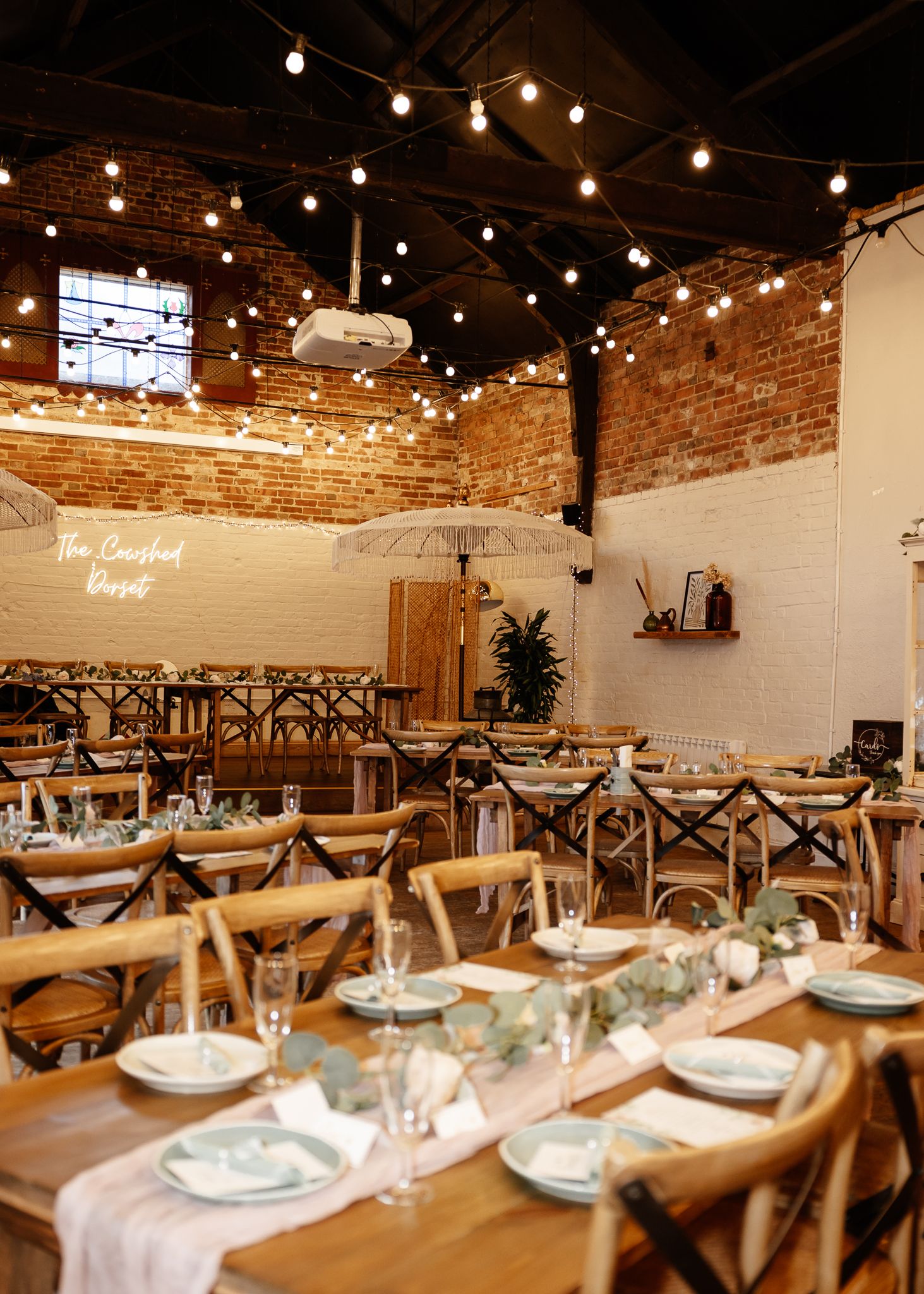 Industrial-chic event space with string lights overhead and long tables set for dining against white and brick walls.