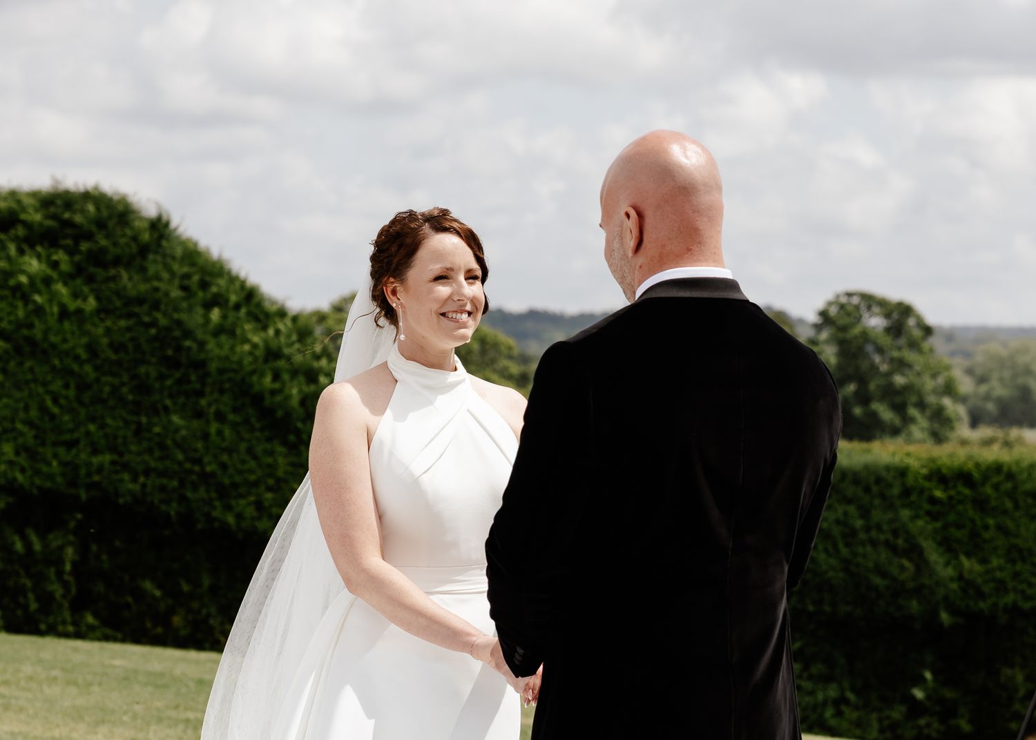 A bride and groom share a moment together outdoors on their wedding day with green hedges and countryside in the background.