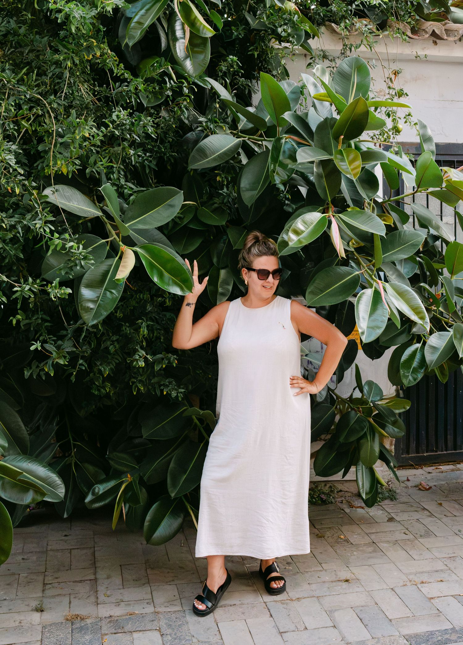 Woman in white midi dress and black sandals posing beside large tropical rubber tree plants outdoors.