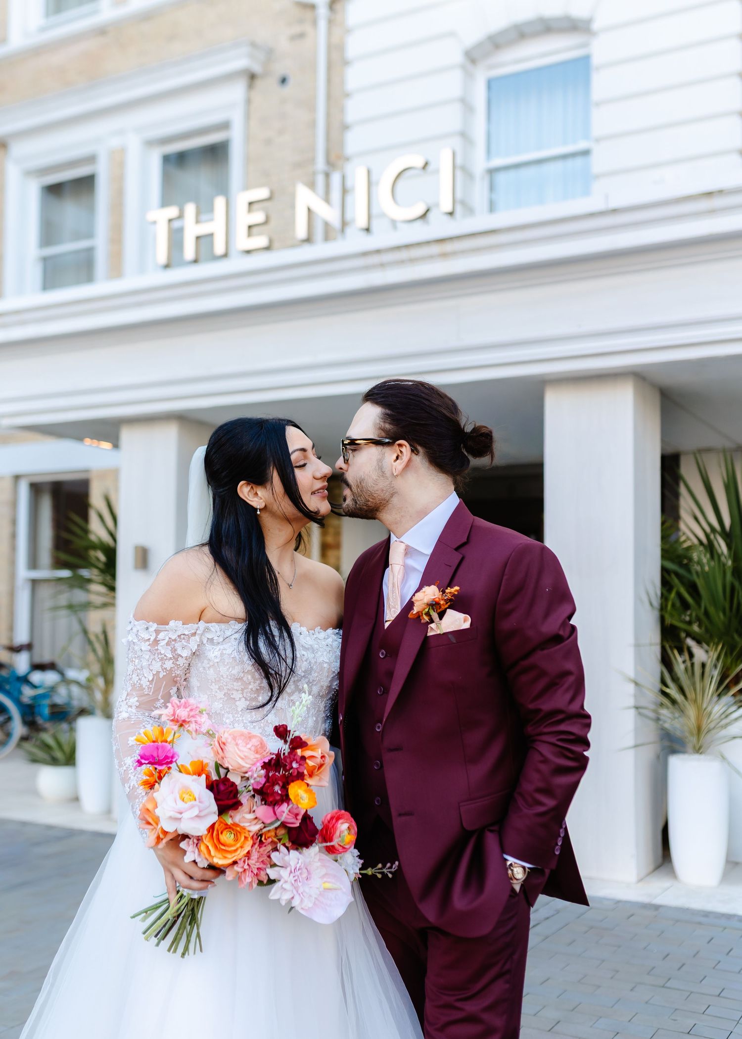 Bride and groom share a romantic moment outside The Nici hotel, bride holding vibrant pink and orange bouquet.