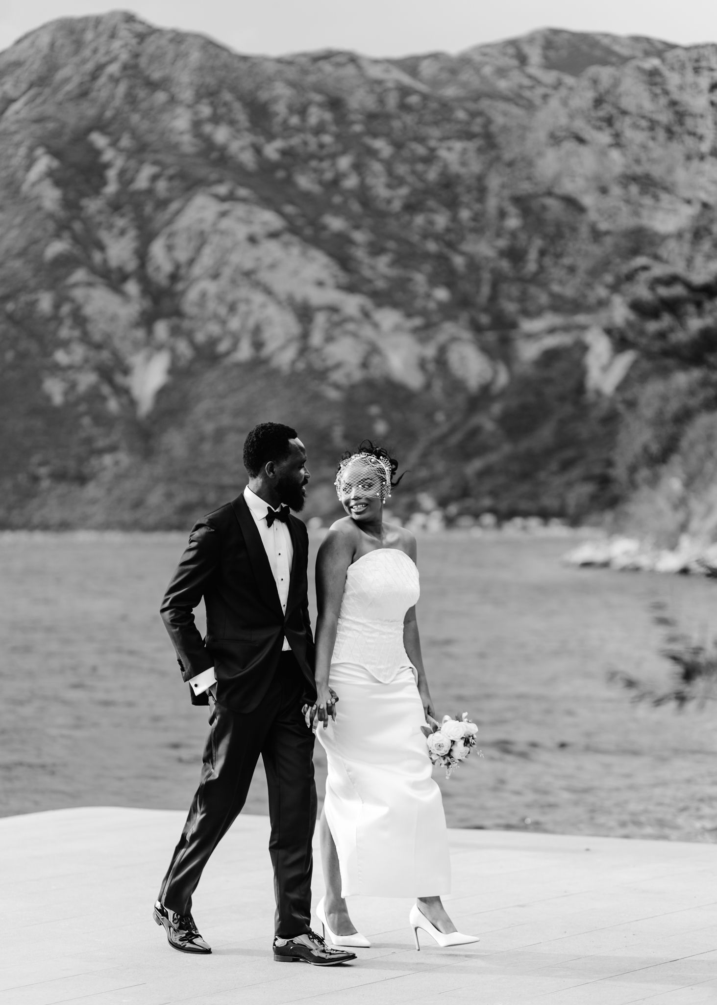 A couple in formal wedding attire walks along a scenic coastal path with rocky mountains in the background.