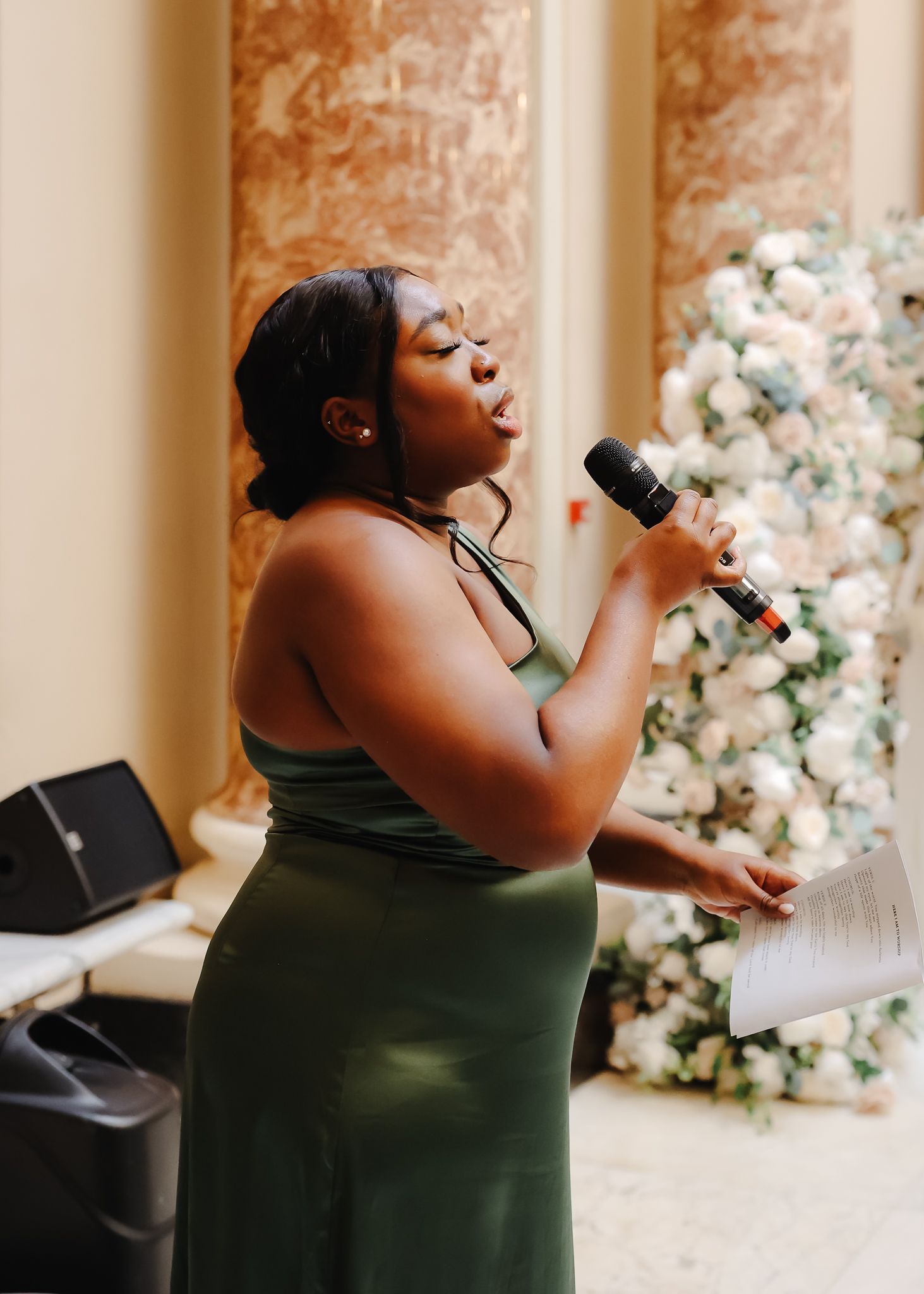 A singer in a dark green sleeveless dress performs passionately with a microphone against a floral backdrop.