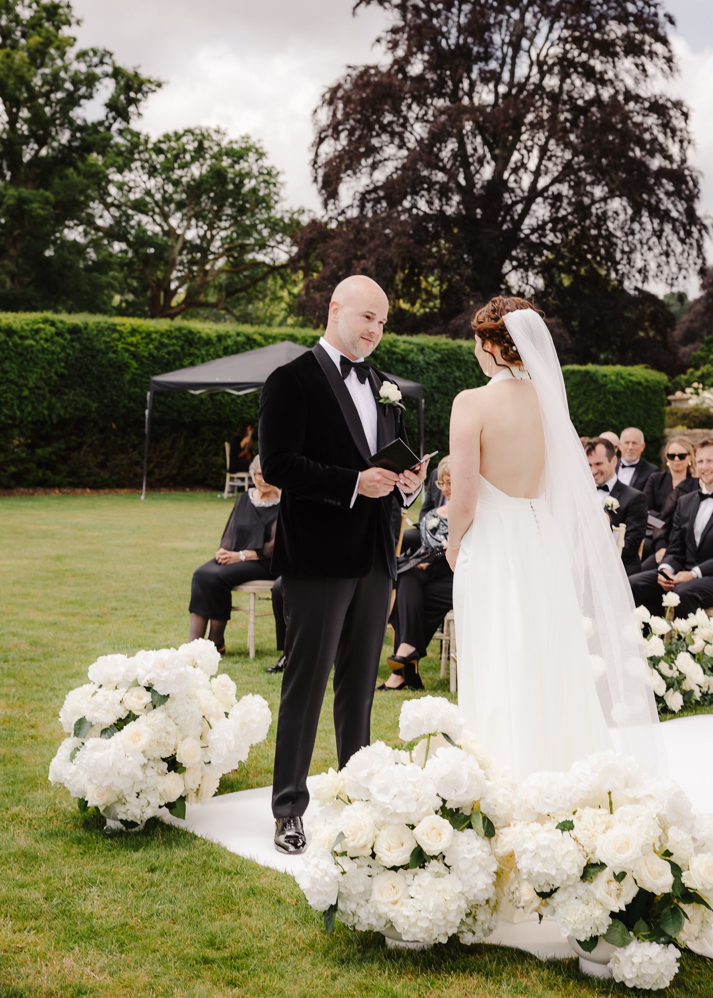 A wedding ceremony sequence showing the couple exchanging vows outdoors with white floral arrangements on a green lawn.