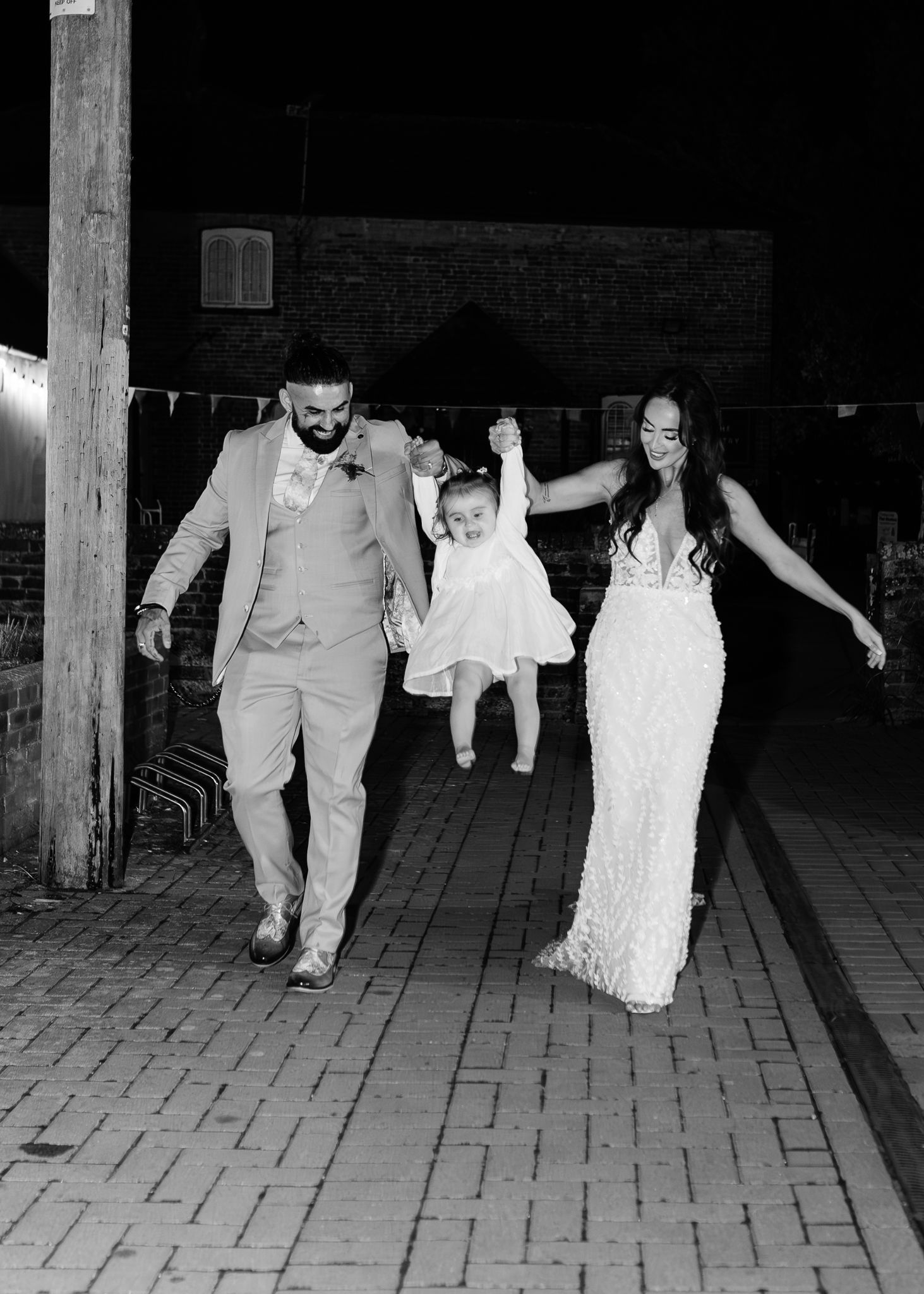 A couple in wedding attire swings a small child in a white dress between them while walking on a brick path.