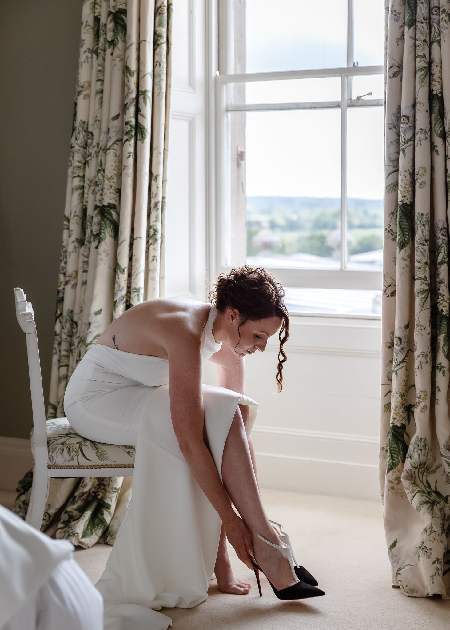 A person in a white dress bends down to adjust their shoe near a window with floral curtains.