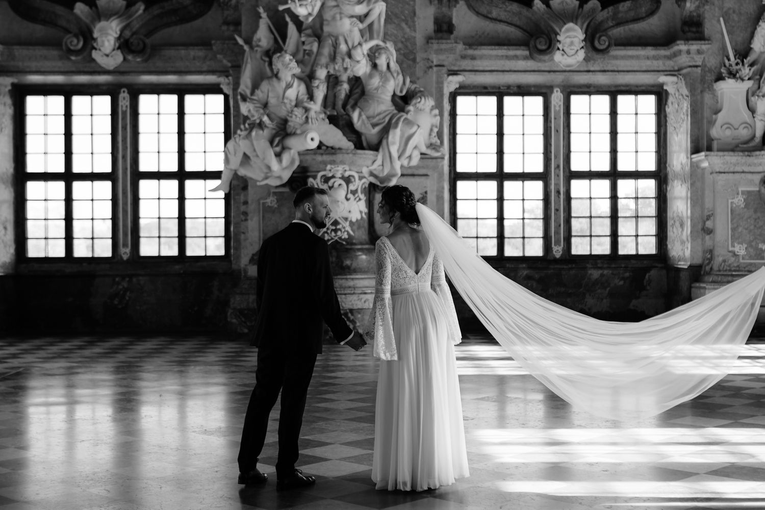 A couple in formal wedding attire dances gracefully in an elegant ballroom with ornate windows and decor.