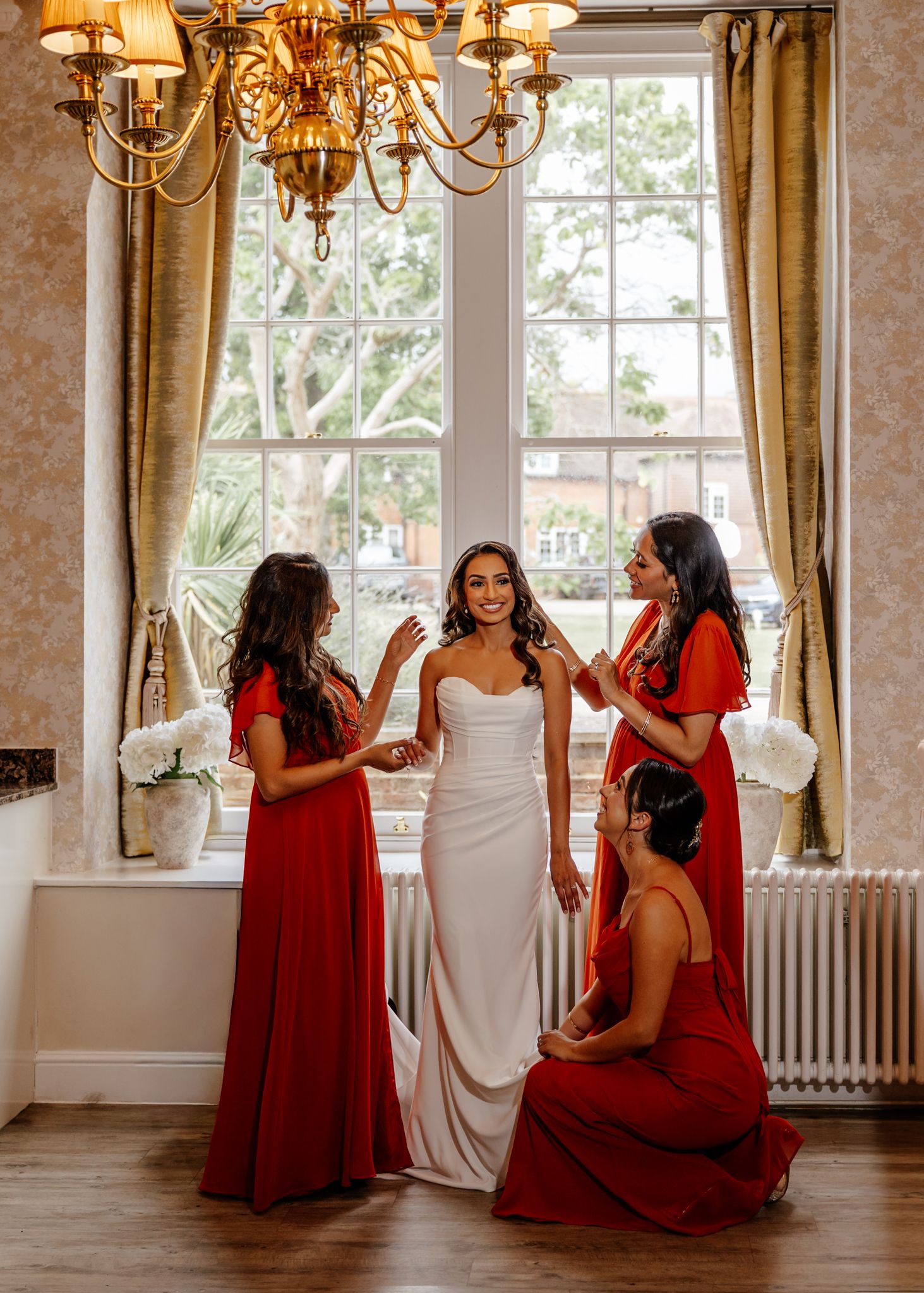 A bridal party in red dresses gathers by a bright window under an elegant chandelier in a classic interior setting.