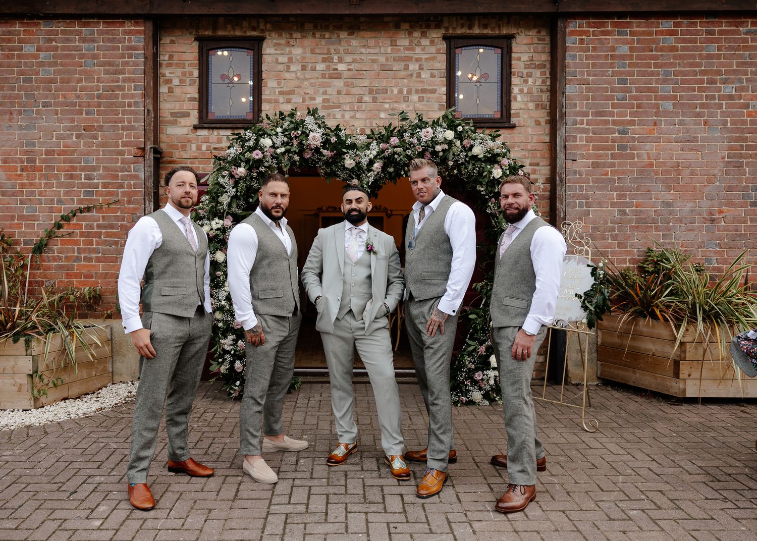 Wedding party members in light gray suits and brown shoes pose in front of a brick wall with climbing vines.
