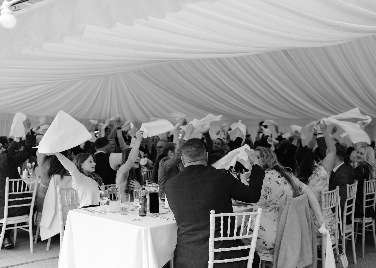 Black and white photo of wedding guests waving napkins in a decorated marquee tent during a celebration.