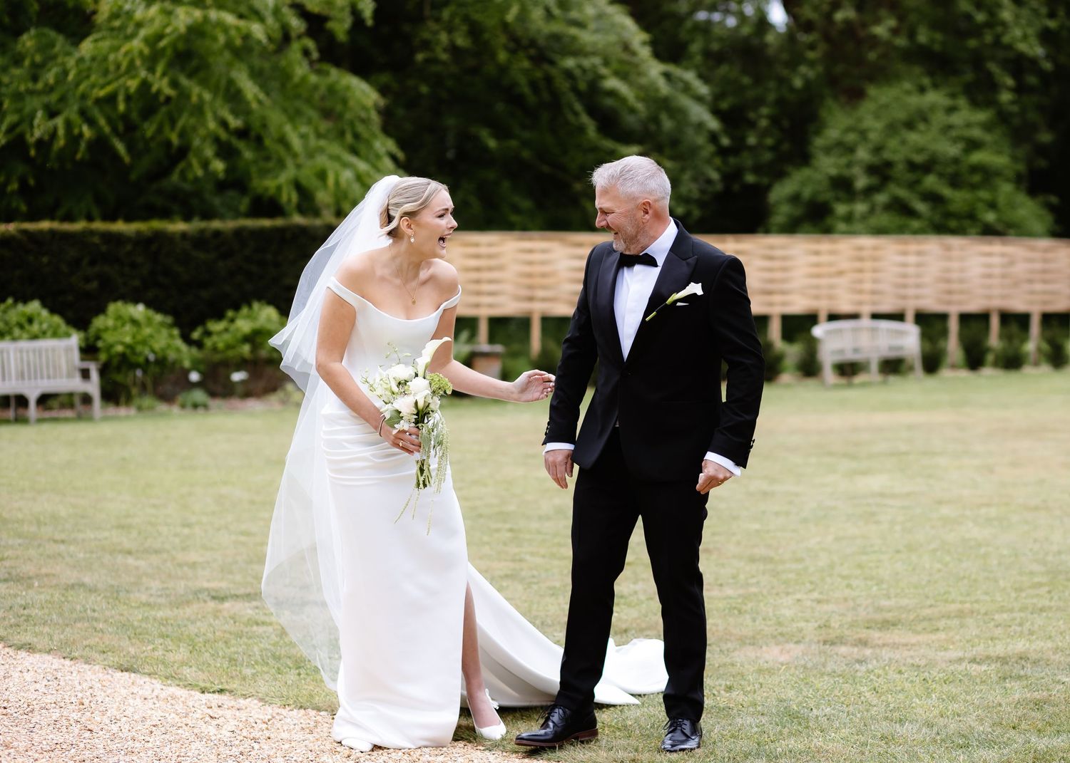 Bride in white gown and groom in black tuxedo holding hands on green lawn at outdoor wedding venue.