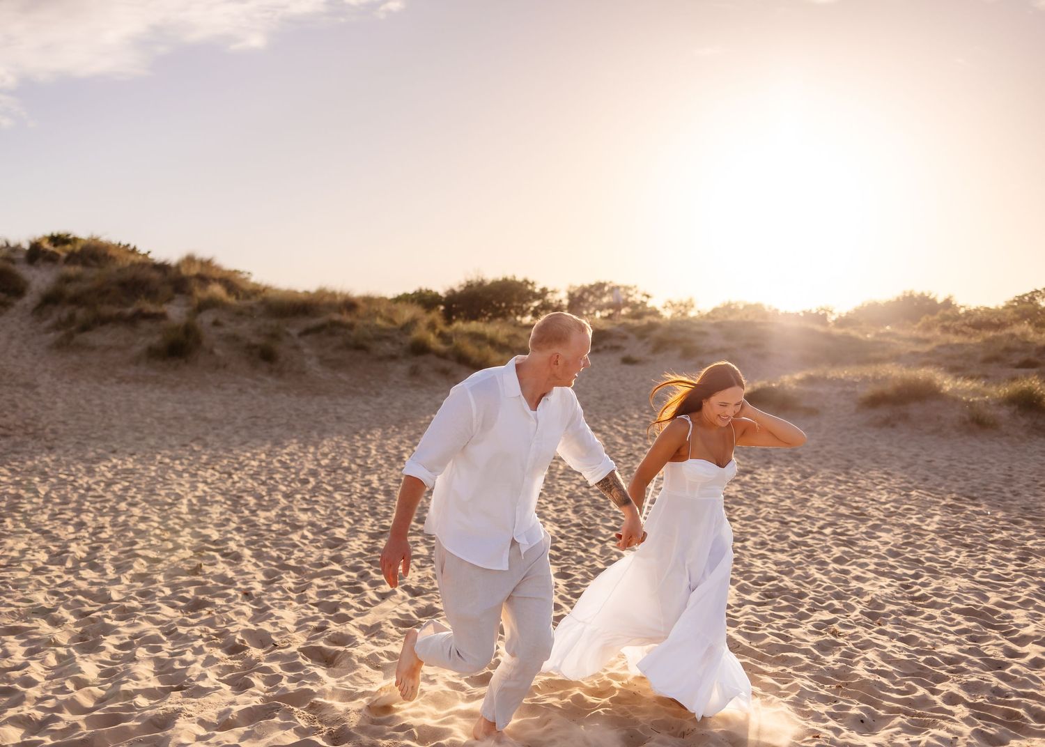 Couple in white outfits holding hands running on sandy beach at golden sunset.