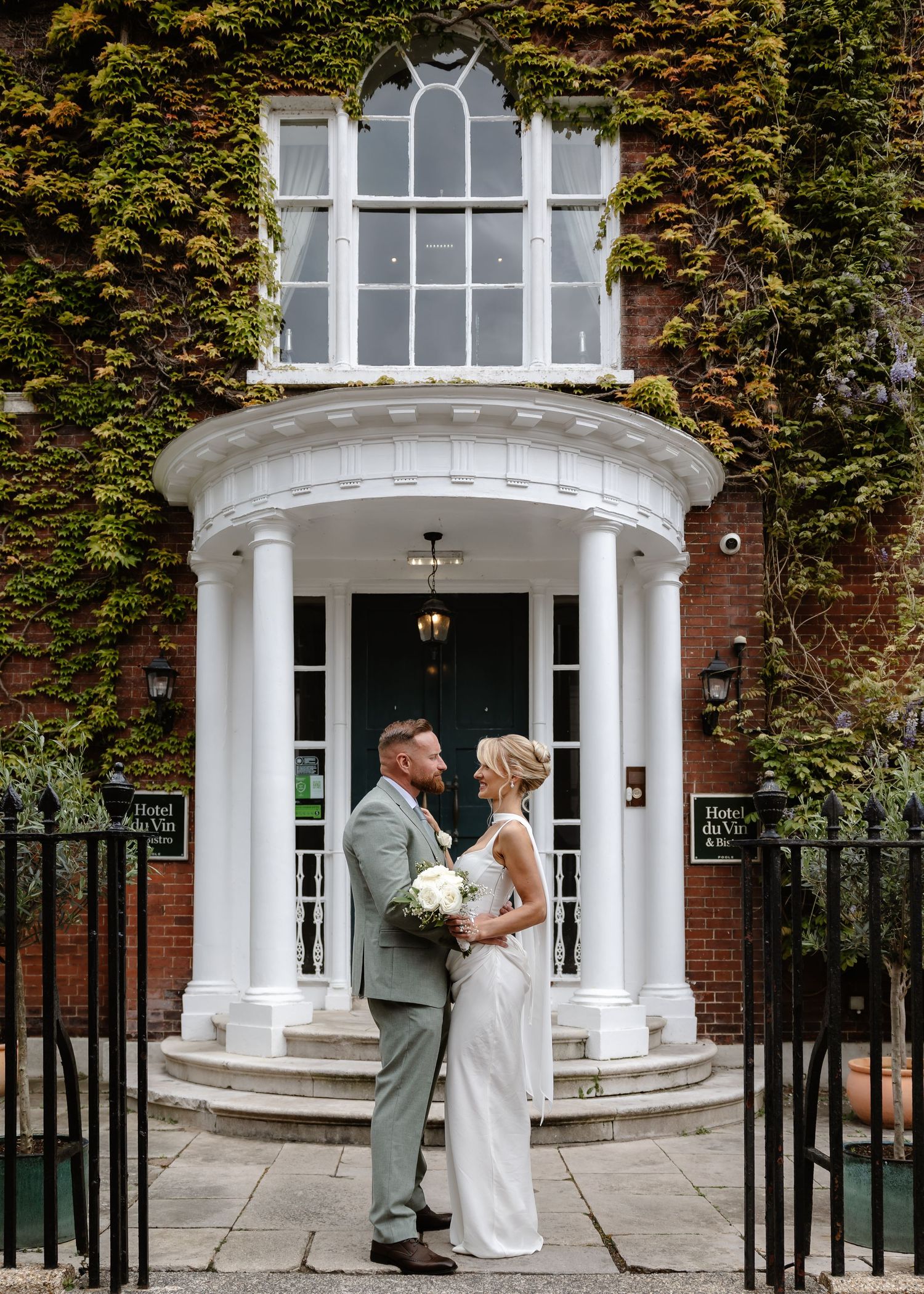 Bride and groom embracing outside ivy-covered Hotel du Vin entrance with white columns on their wedding day.