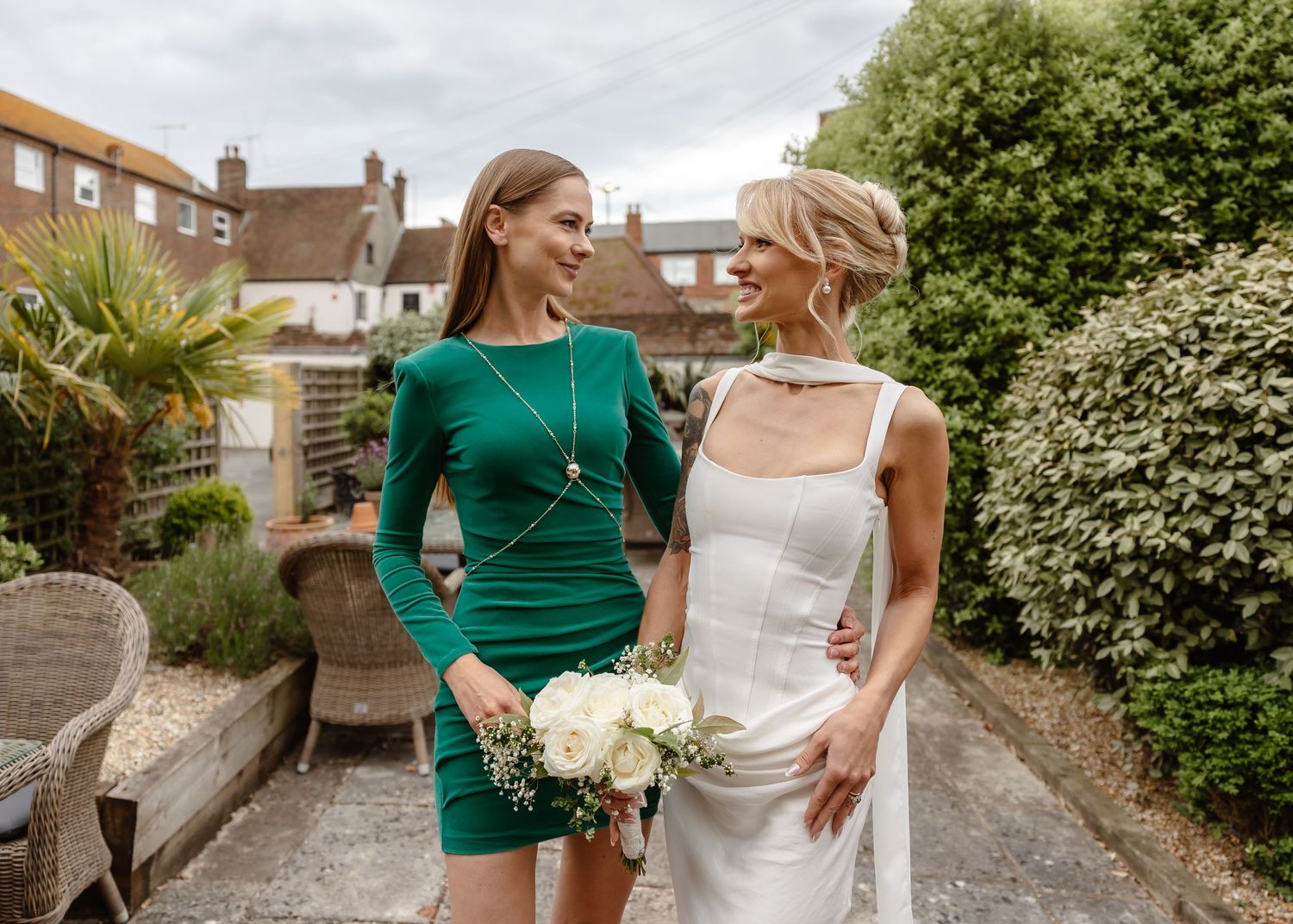 Bride in white dress and bridesmaid in green dress holding white bouquet outdoors at a garden wedding.