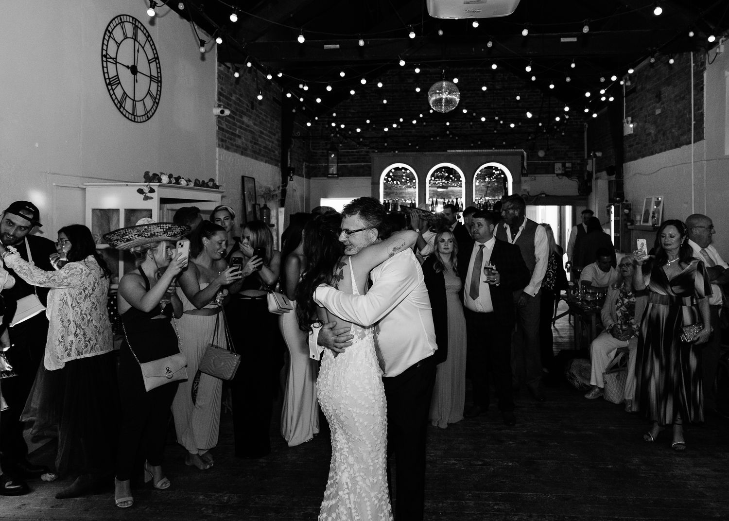 Black and white photo of wedding guests dancing under twinkling lights in a rustic venue.