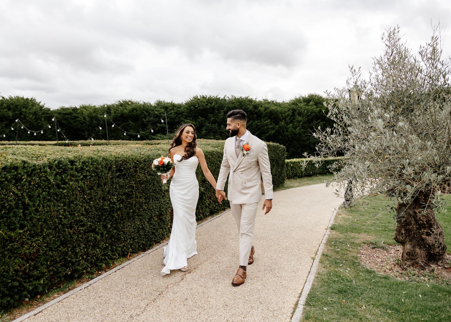 A couple walks hand in hand down a scenic gravel path lined with lush hedges and olive trees on a sunny day.