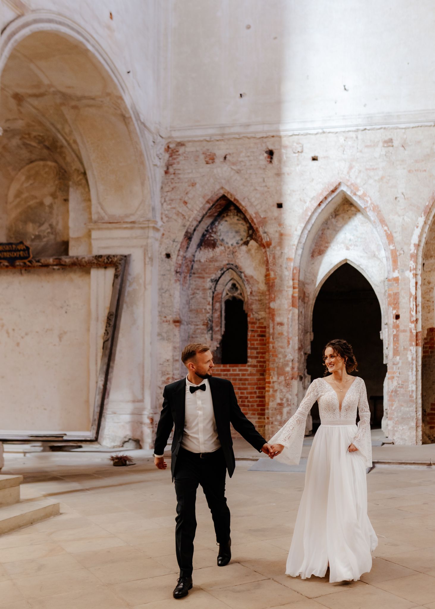 Couple in wedding attire strolling through historic brick and stone archways.