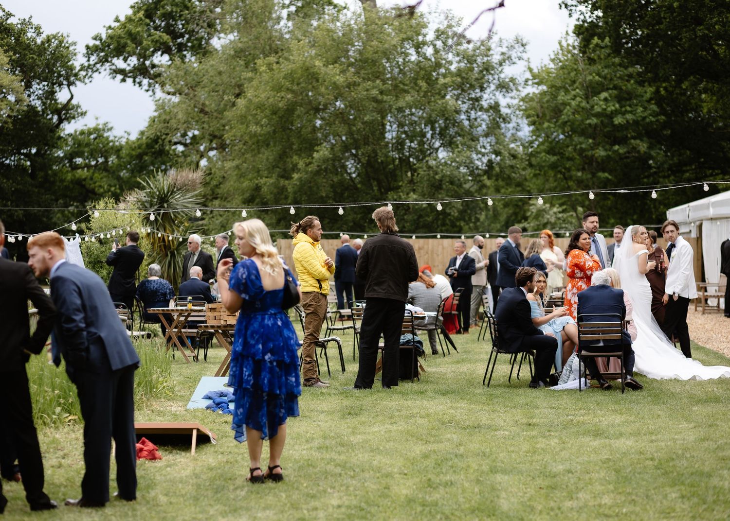Outdoor wedding reception on a sunny day with guests mingling on a green lawn, string lights, and a bride in a white dress.