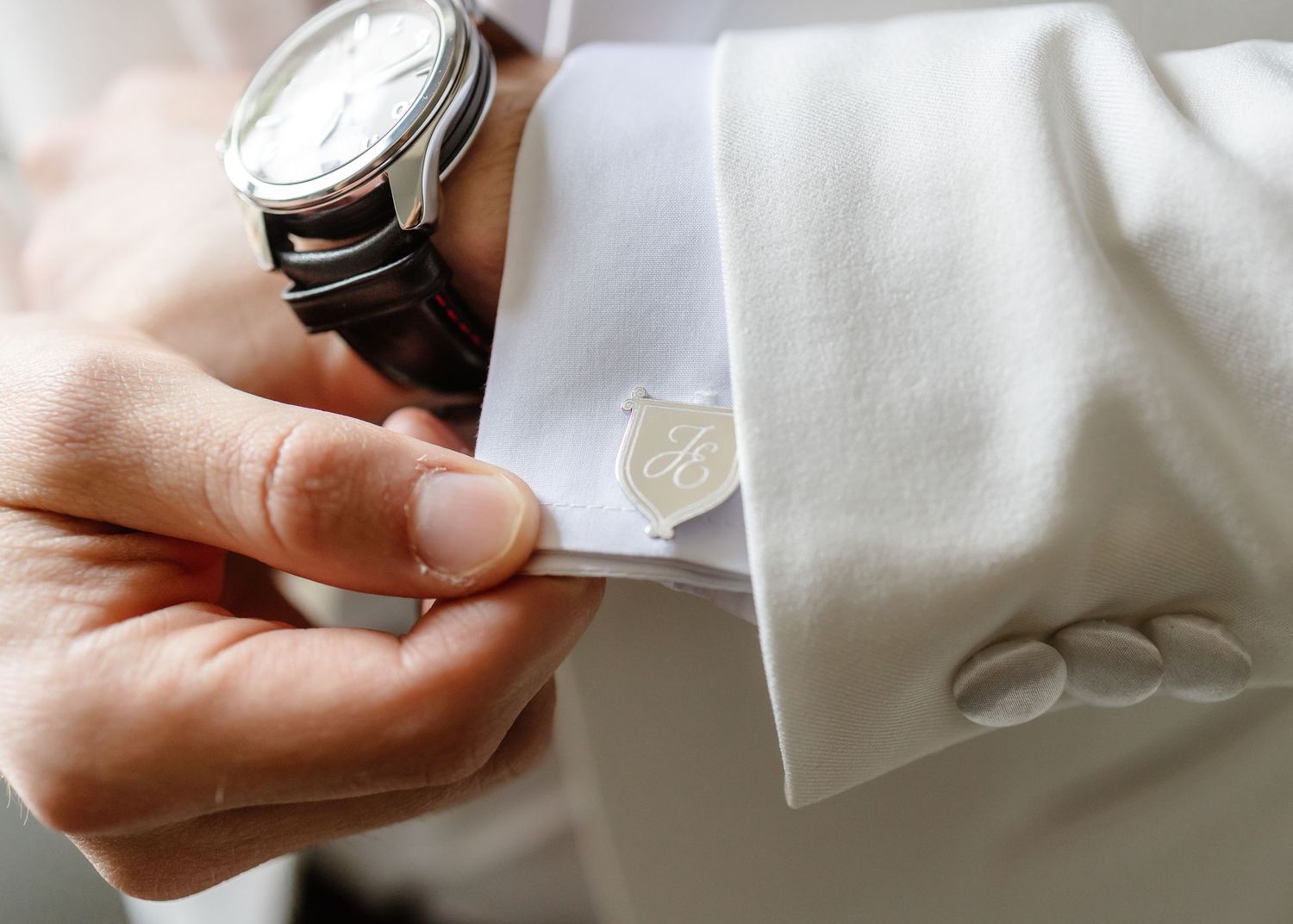 Man in white tuxedo jacket adjusting engraved silver cufflink while wearing a classic brown leather strap wristwatch.