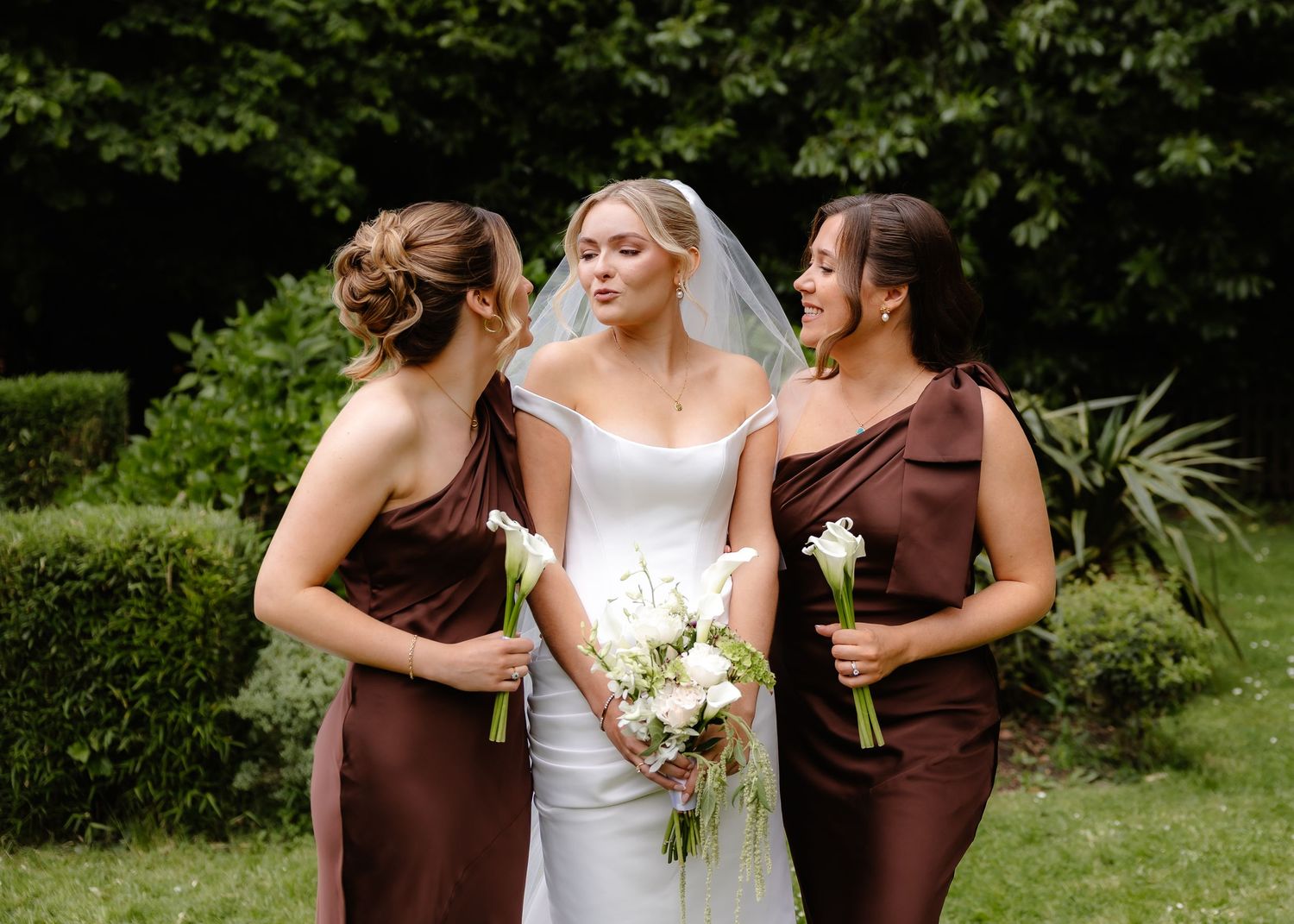 Bride in white gown with veil flanked by two bridesmaids in brown satin dresses holding bouquets outdoors.