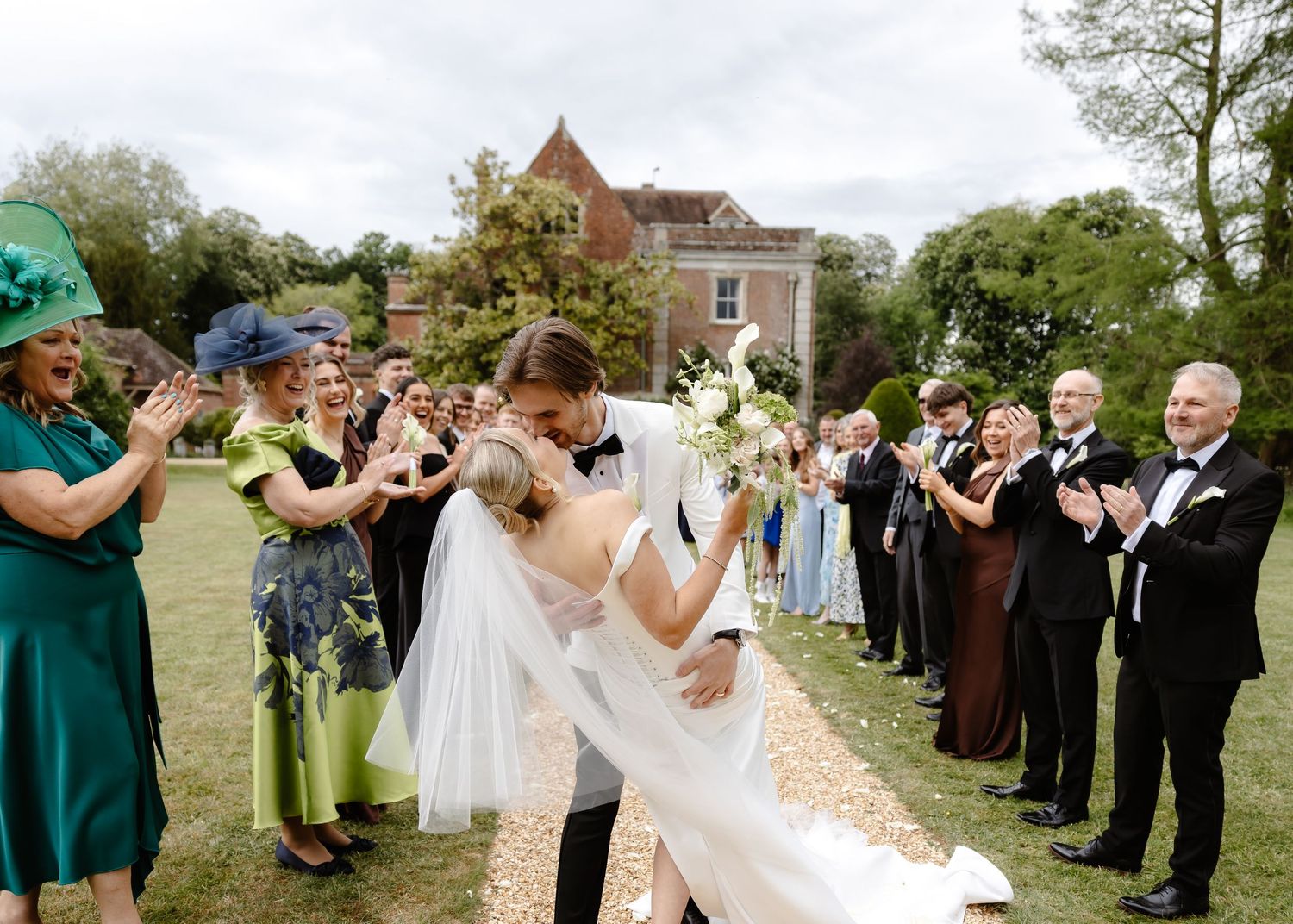 Bride and groom share a romantic kiss outdoors at a wedding celebration with guests applauding beside a historic manor.