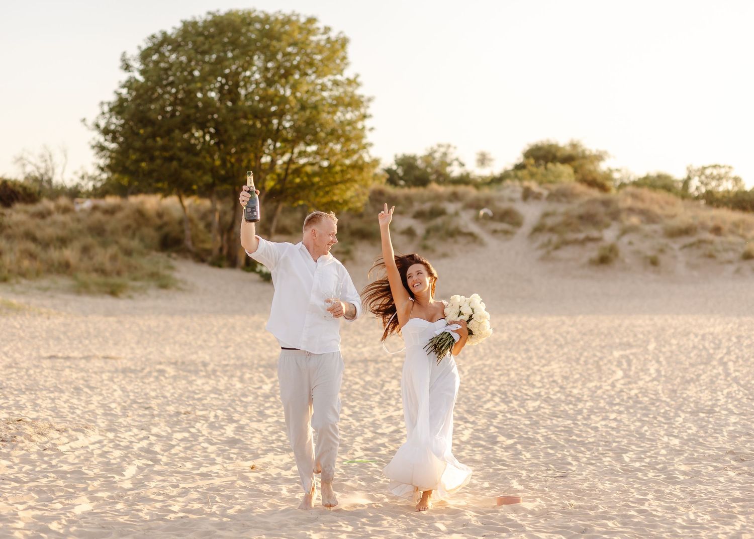Joyful newlyweds celebrating on a sandy beach, bride holding bouquet and groom raising champagne bottle at sunset.