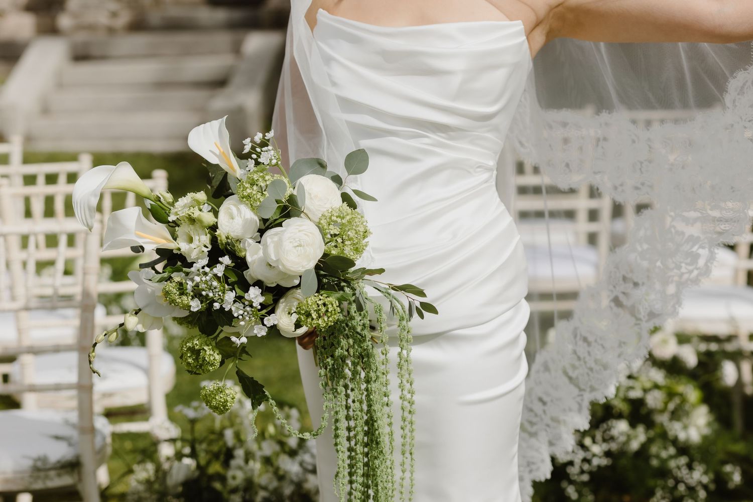 Bride in white gown holding elegant green and white wedding bouquet with cascading vines at outdoor ceremony.