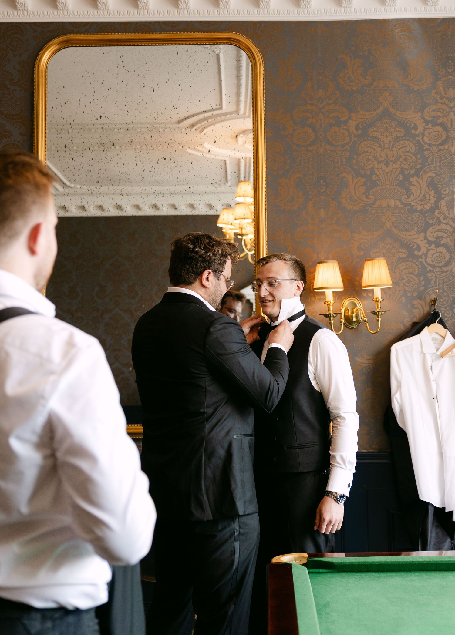 Groomsmen helping each other with bow ties in an elegant room with gold mirror and damask wallpaper.