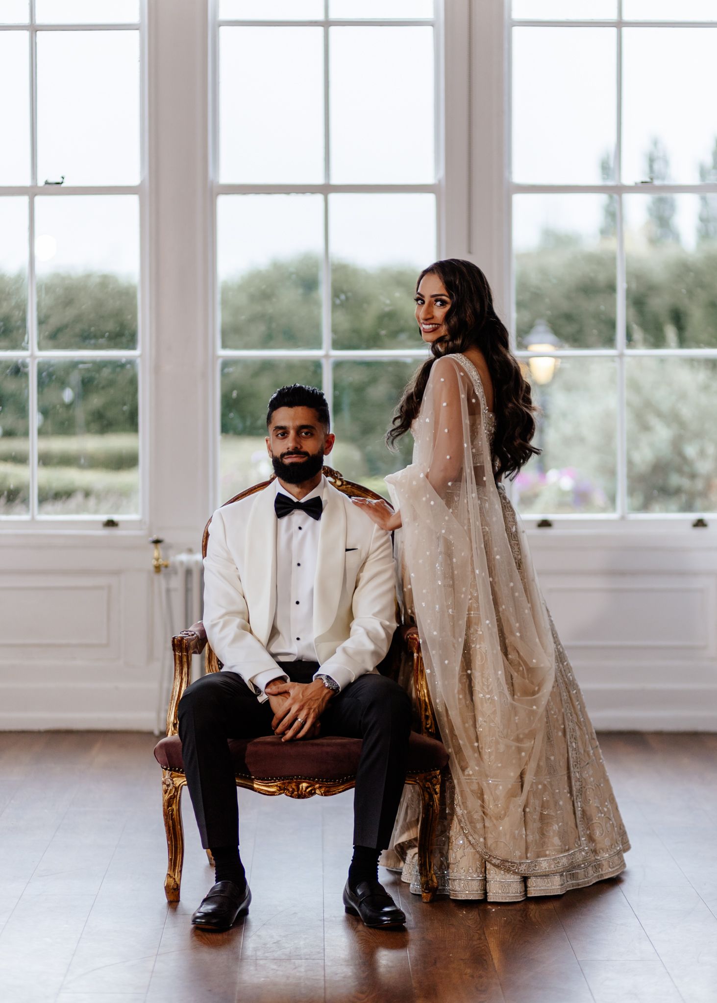 A wedding couple sits on chairs in an elegant white room with large windows, creating a romantic atmosphere.