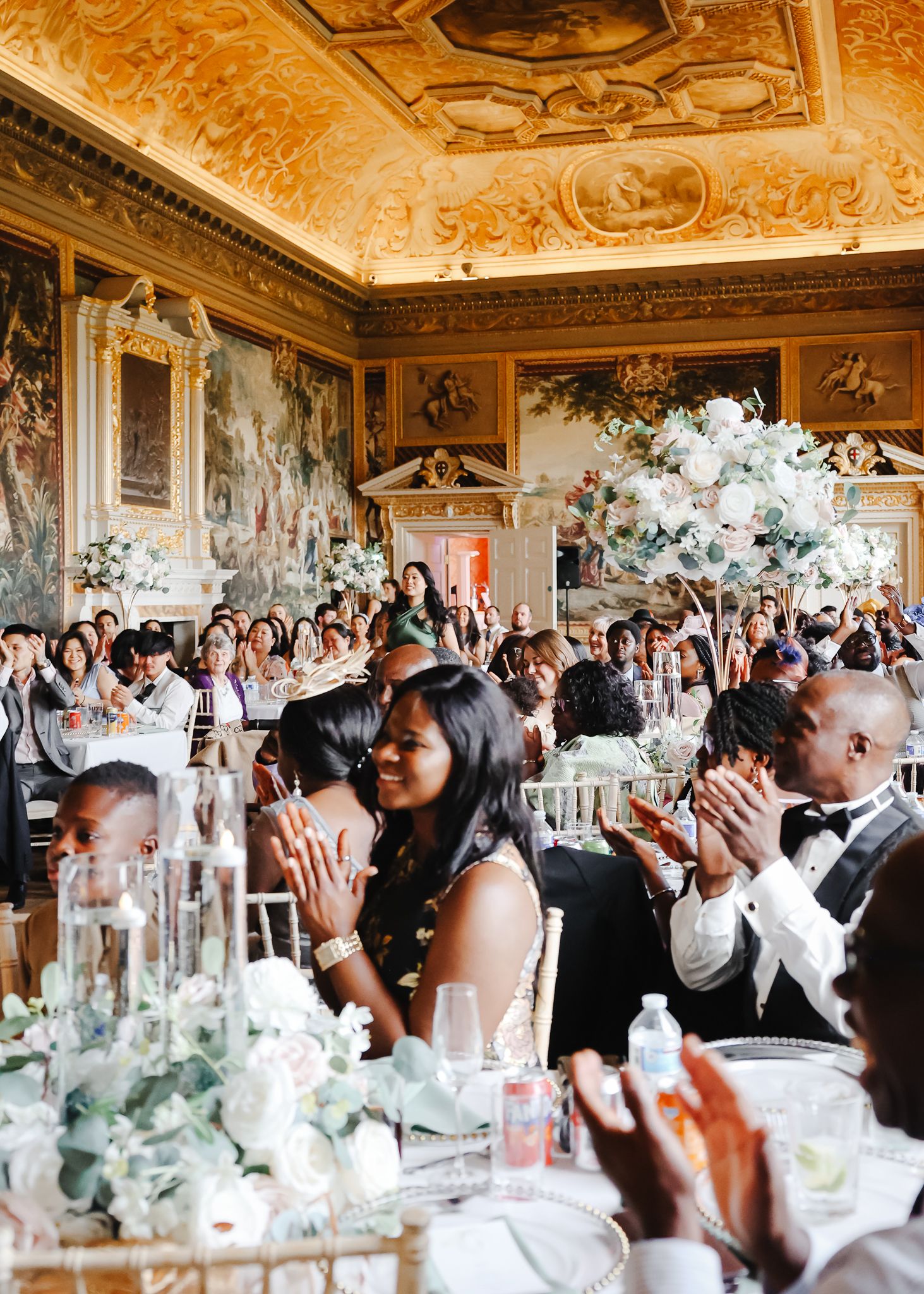 Wedding guests raise their hands in celebration at an elegant reception with ornate ceiling and floral centerpieces.